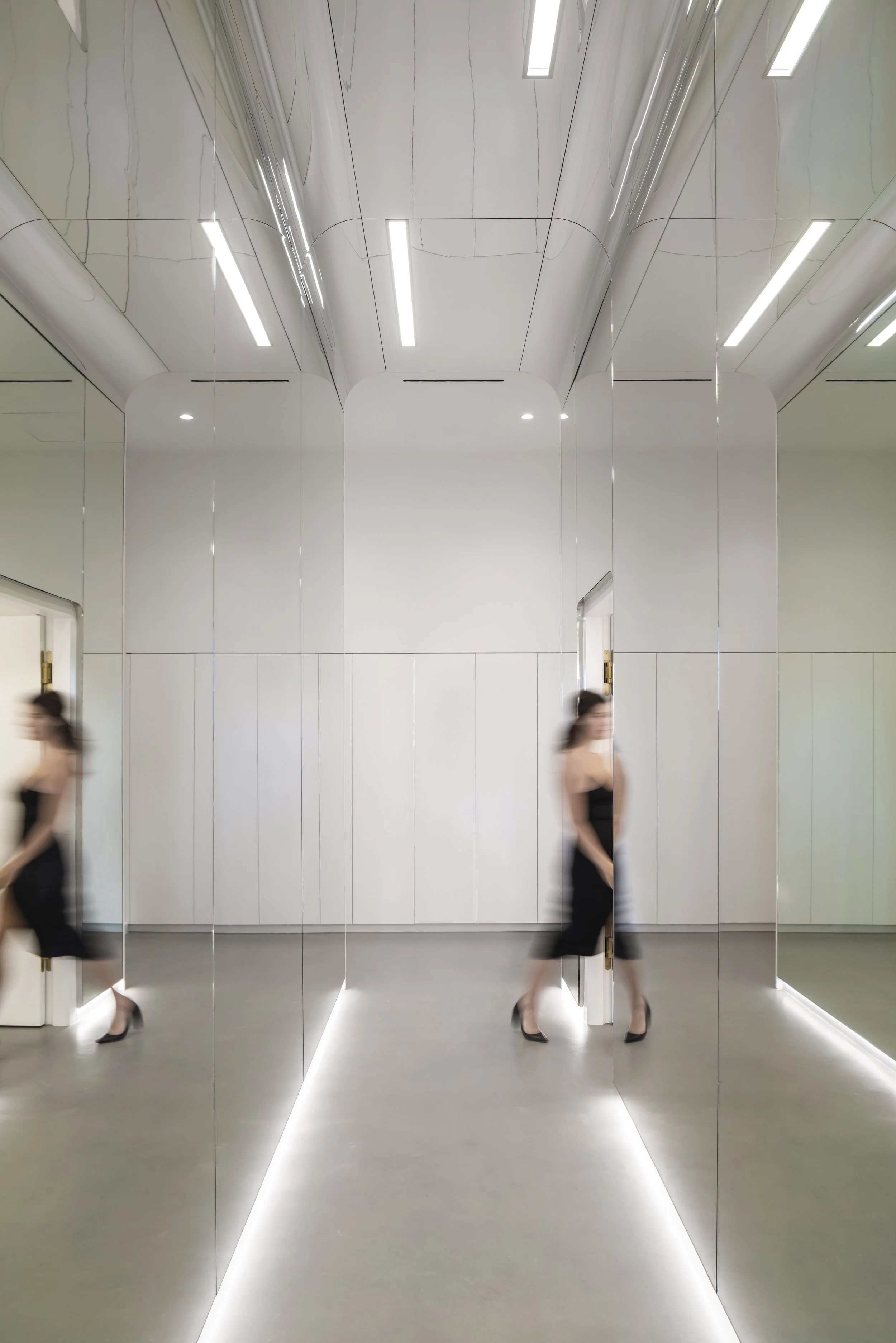 Blurred woman walking in minimalist, modern room with reflective surfaces and overhead lighting.