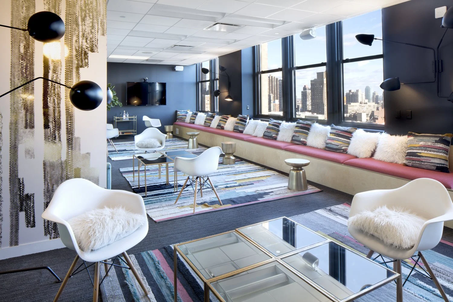 Bright modern lounge with colorful seating, white chairs, patterned rugs, and decorative pillows; large windows with city skyline view.