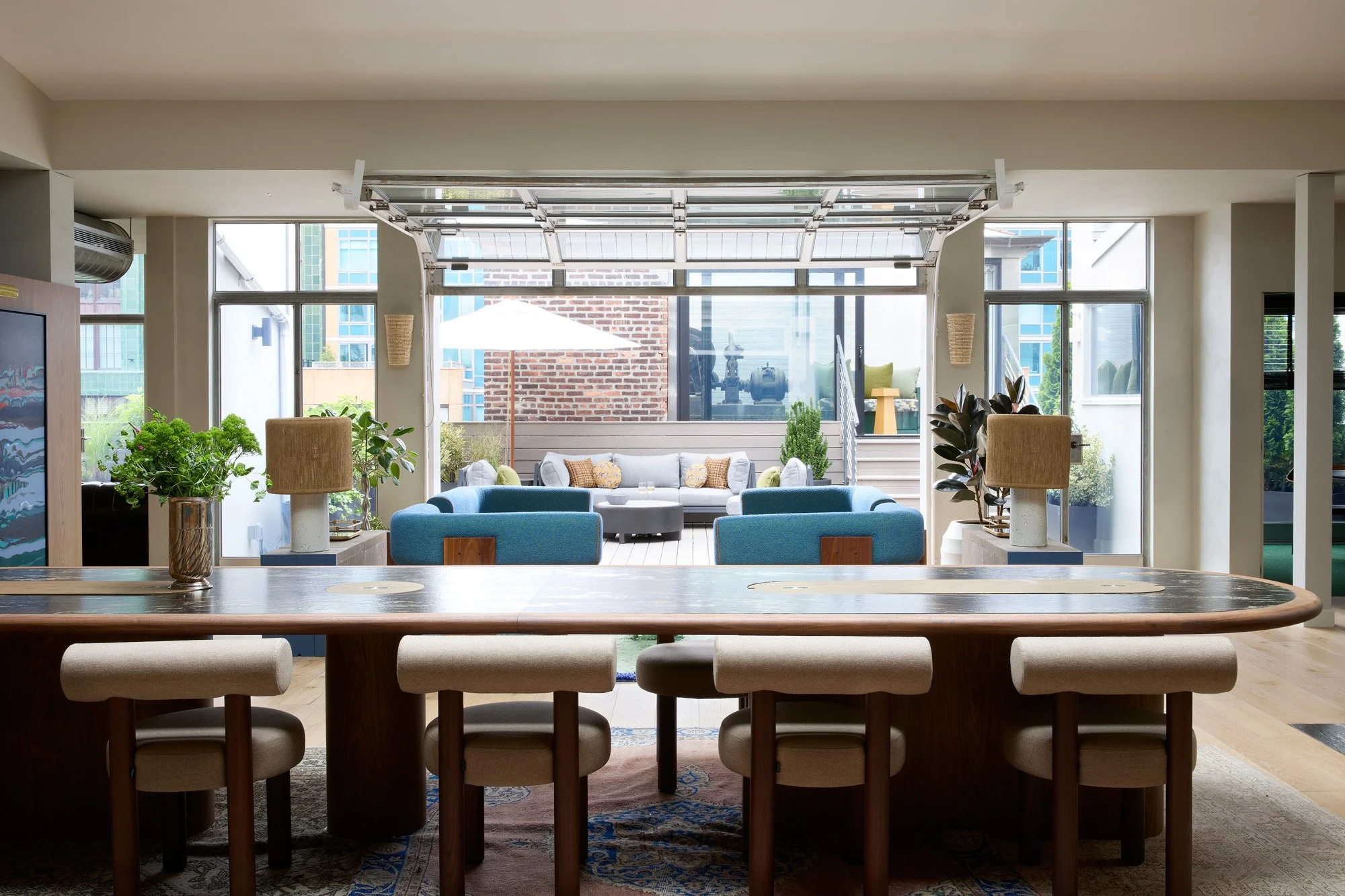 Interior view of a contemporary living space with a large wooden dining table, beige chairs, and a sitting area with blue and gray sofas and an outdoor patio with plants and an umbrella.