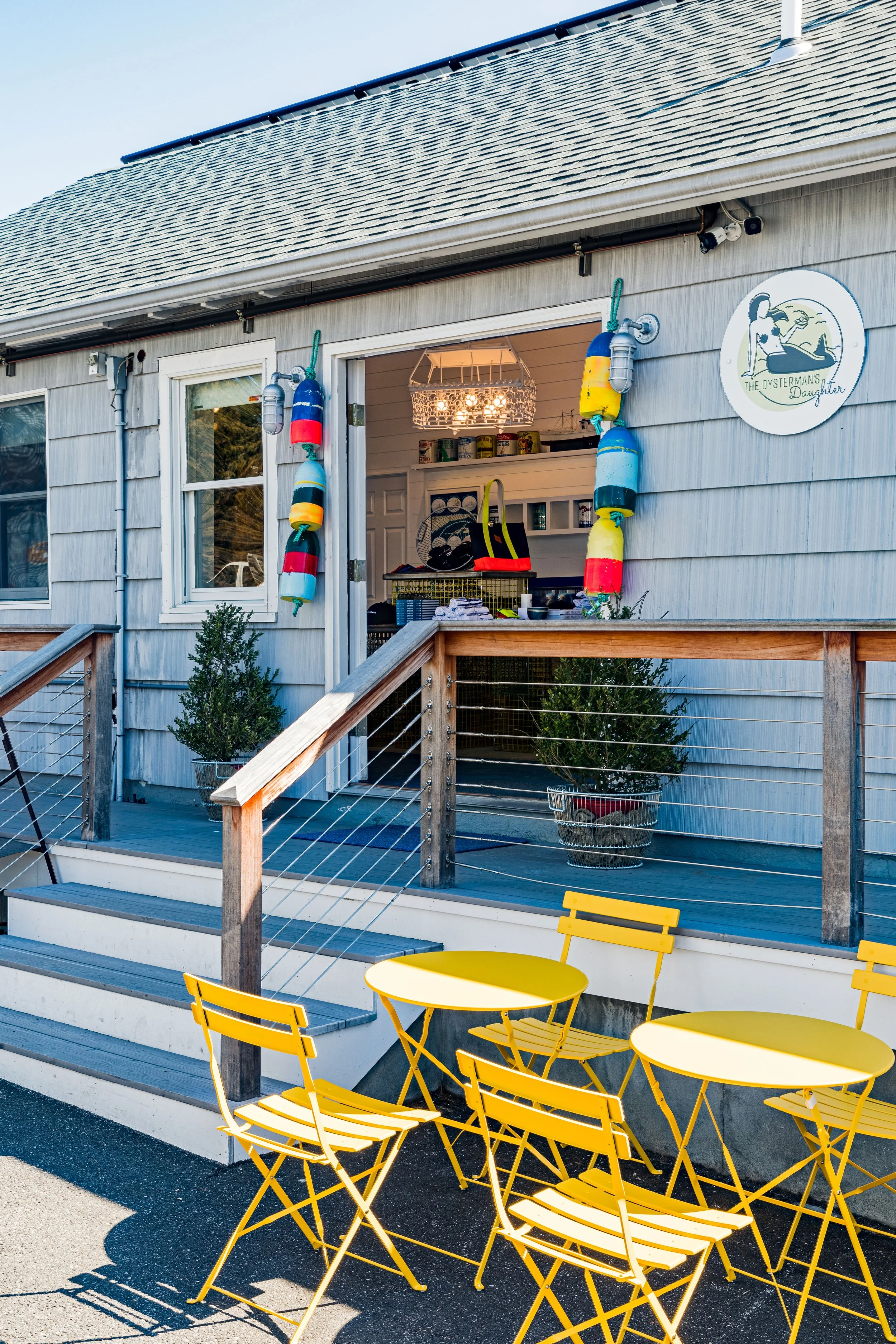 Exterior of a charming cafe with blue siding, colorful buoys, and a sign labeled 'The Oysterman's Daughter.' There's a small porch with railings and yellow outdoor tables and chairs on the pavement.