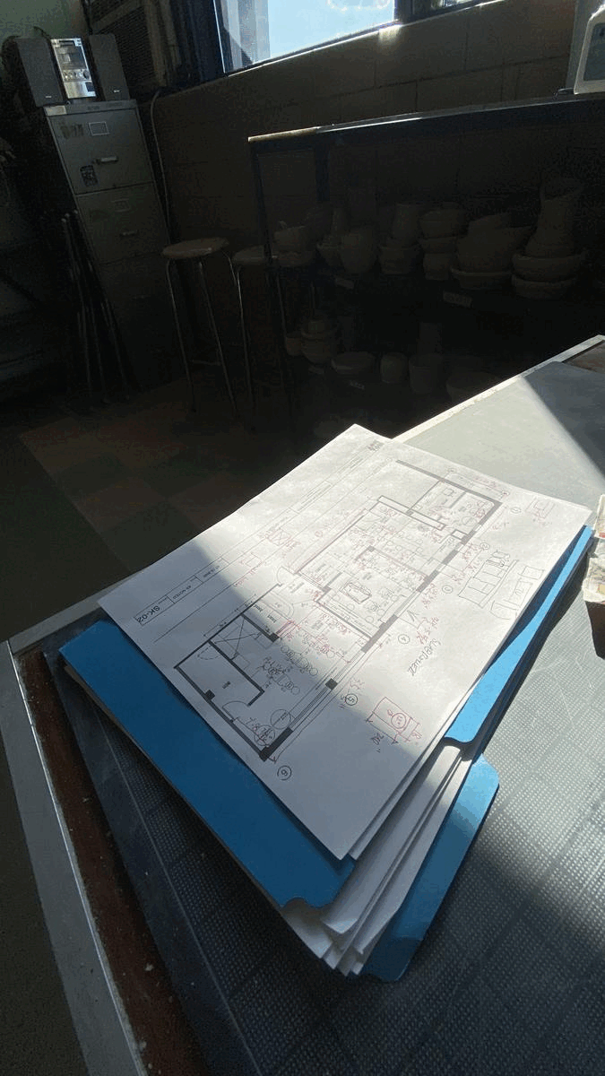 Stack of architectural blueprints on a table under window light, with pottery on shelves in the background.