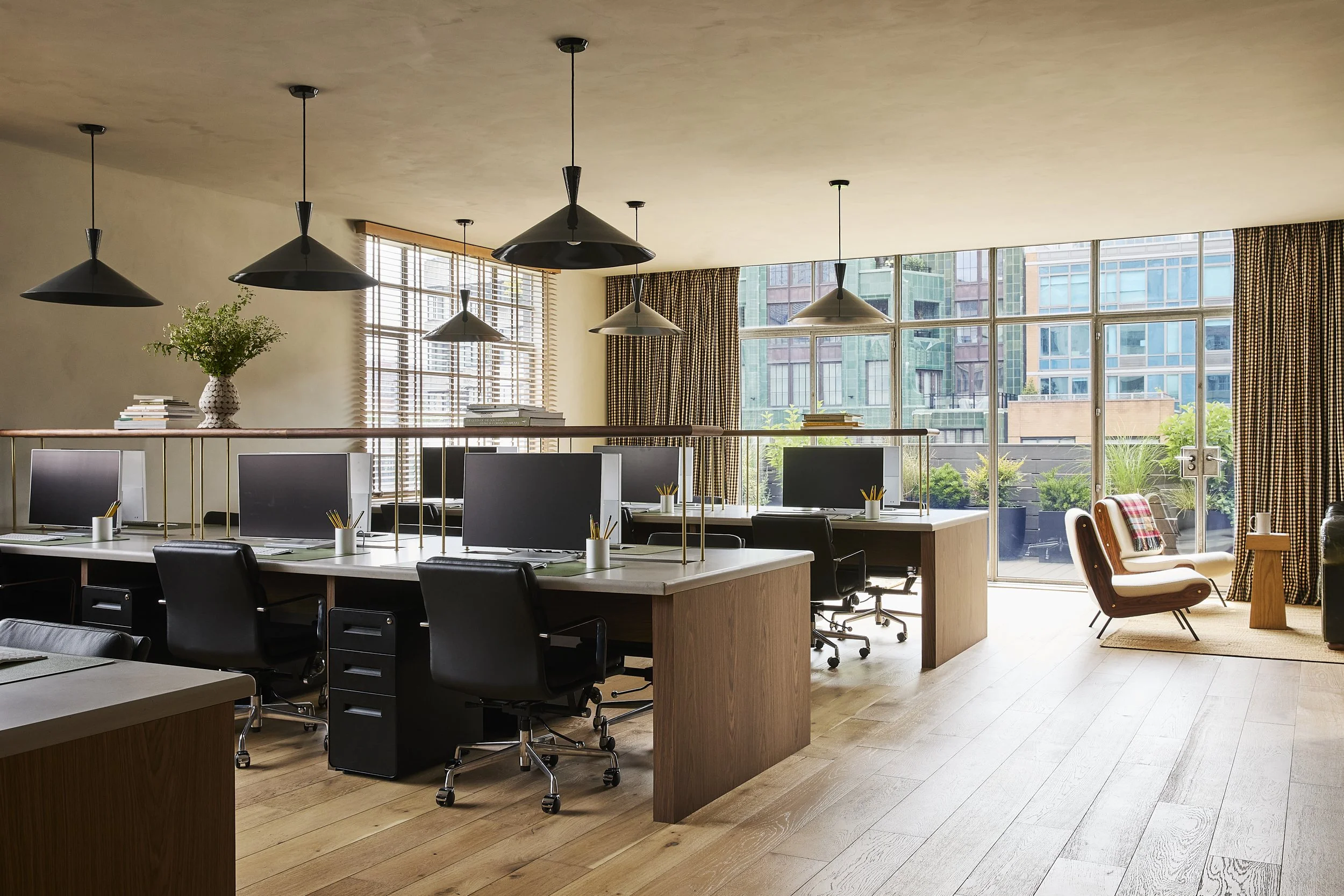 Modern office space with four desks, black chairs, computer monitors, a sitting area with two armchairs, large windows with city buildings outside, wooden floors, ceiling pendant lights, and curtains.
