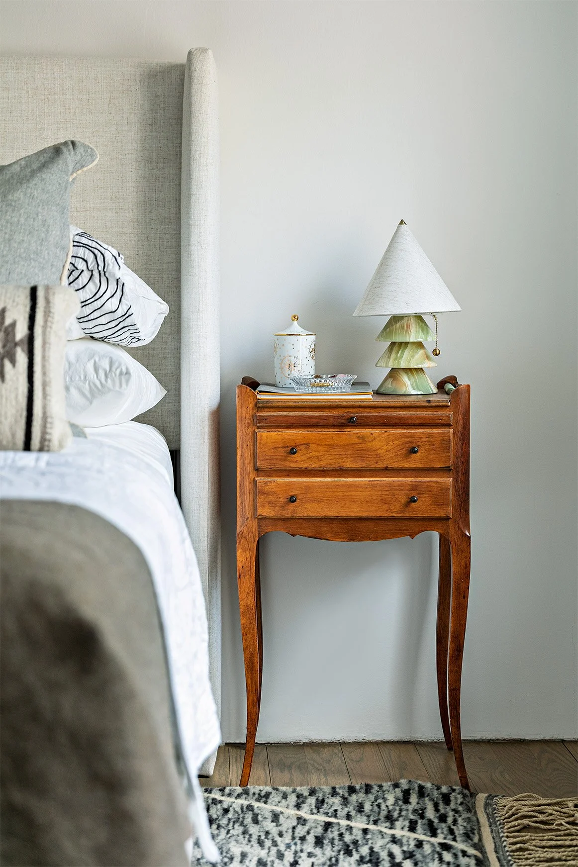 A wooden bedside table with three drawers, a lamp with a tiered stone base and a conical shade, a small decorative container, and some papers, next to a bed with white and striped pillows, in a bedroom with white walls and wooden flooring.