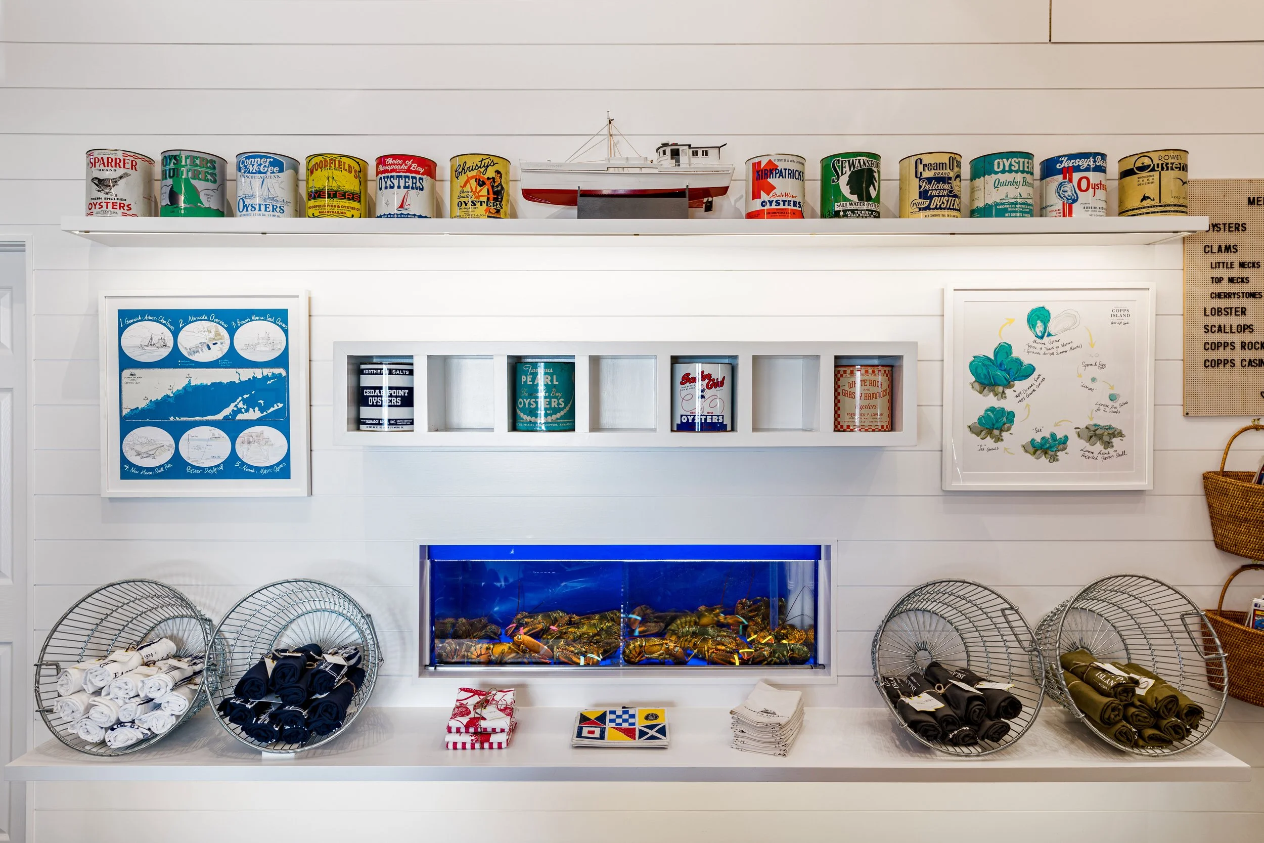 Interior of a seafood-themed store with vintage oyster cans, a model boat, framed nautical maps, lobster tank, and cloth items in baskets.