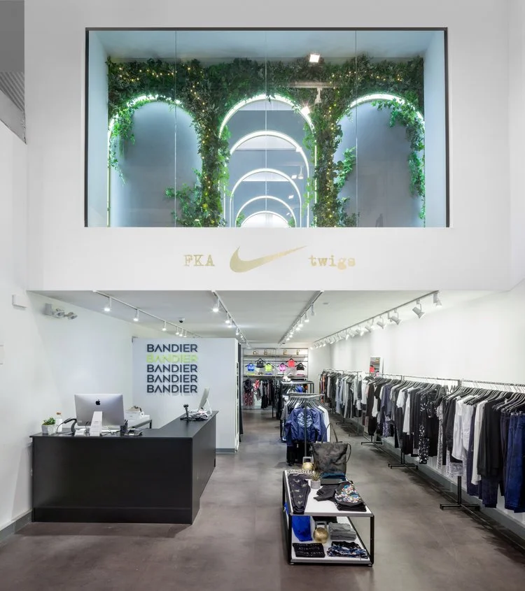 Interior of a retail clothing store with a modern design, featuring racks of apparel on both sides, a central display table with folded clothes, and a checkout counter with a computer. Above, a decorative alcove with vibrant greenery and illuminated rings is visible, with signage displaying 'FKA twigs' and a Nike logo.