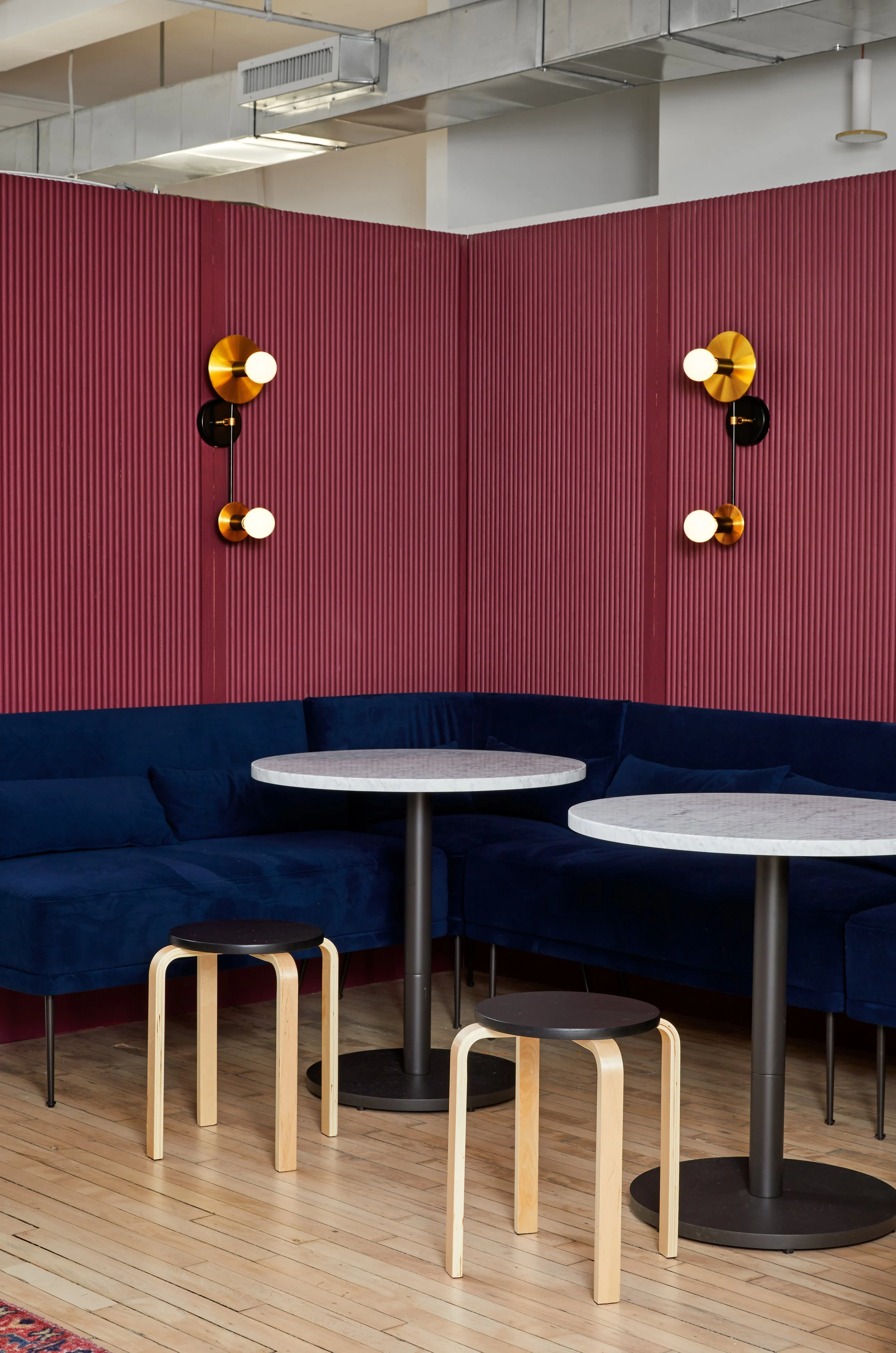 Modern seating area with dark blue velvet sofa, round marble tables, light wood stools, and gold wall sconces against a burgundy wall panel. Hardwood floor and industrial ceiling visible.