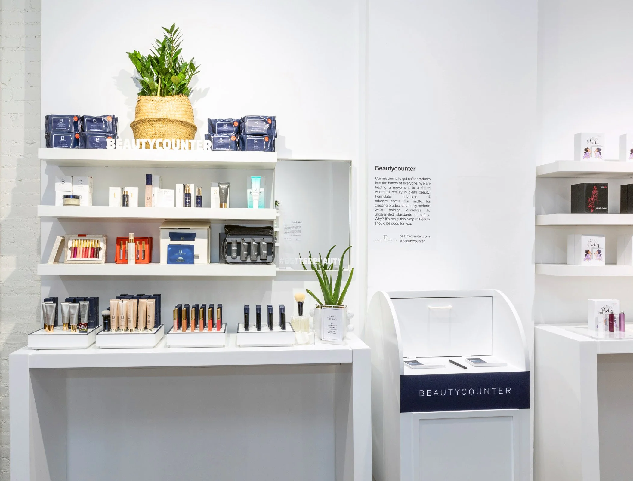 Retail display of skincare and makeup products with "Beautycounter" branding, featuring makeup items on shelves, a potted plant, and informational signage.