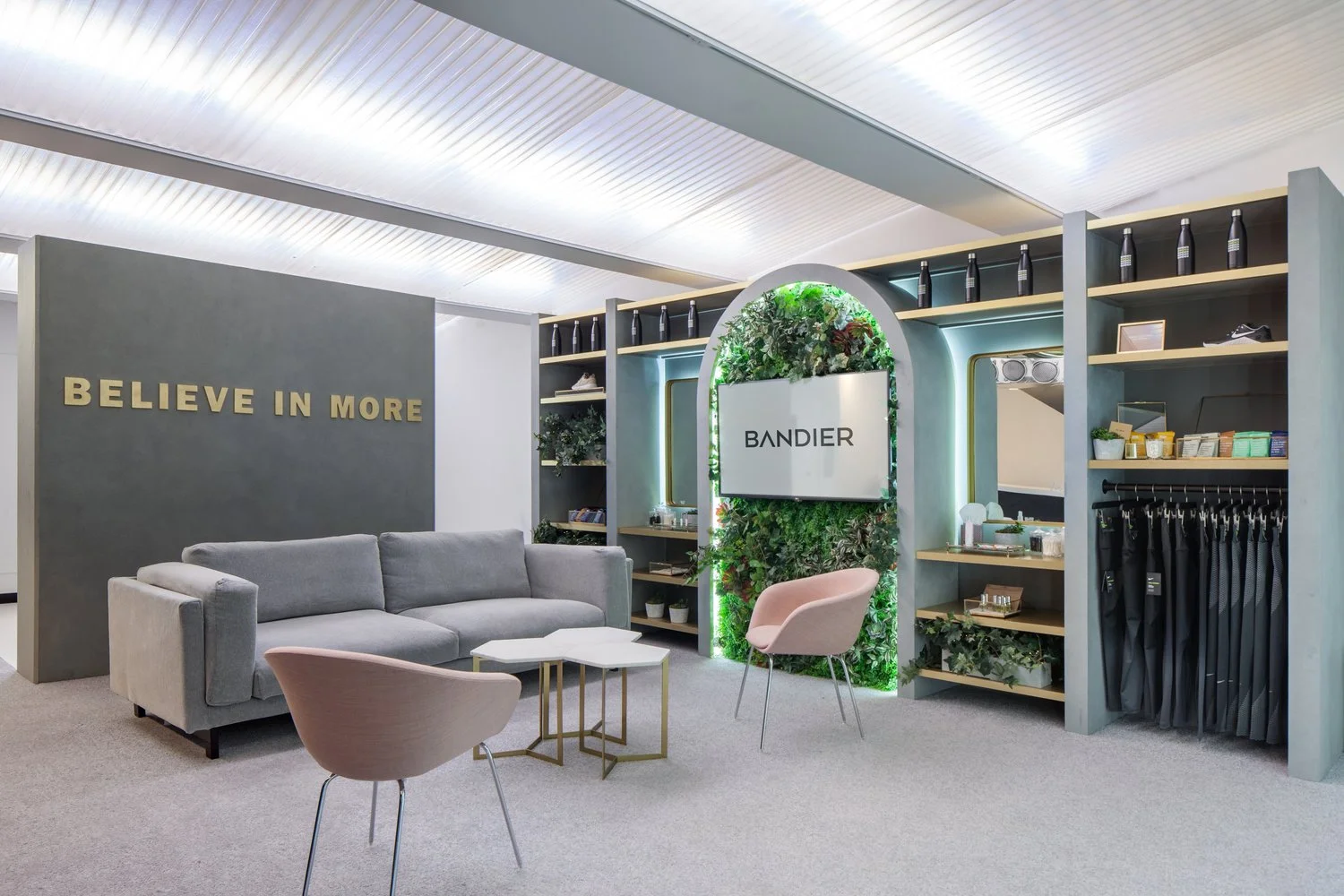 Modern retail lounge with a gray sofa, pink chairs, and shelves displaying products. A sign reads 'Believe in More' on a wall, and a green wall with the name 'Bandier' is featured.