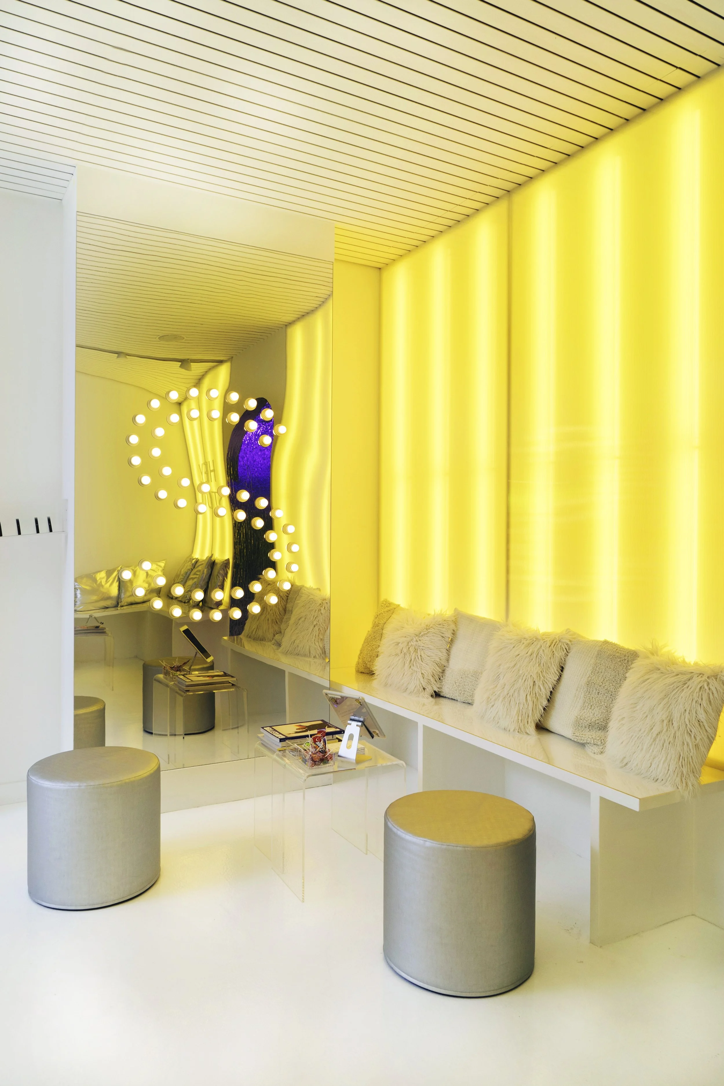 Modern lounge area with yellow illuminated walls, round silver stools, a bench with fluffy pillows, and acrylic tables with magazines and devices.