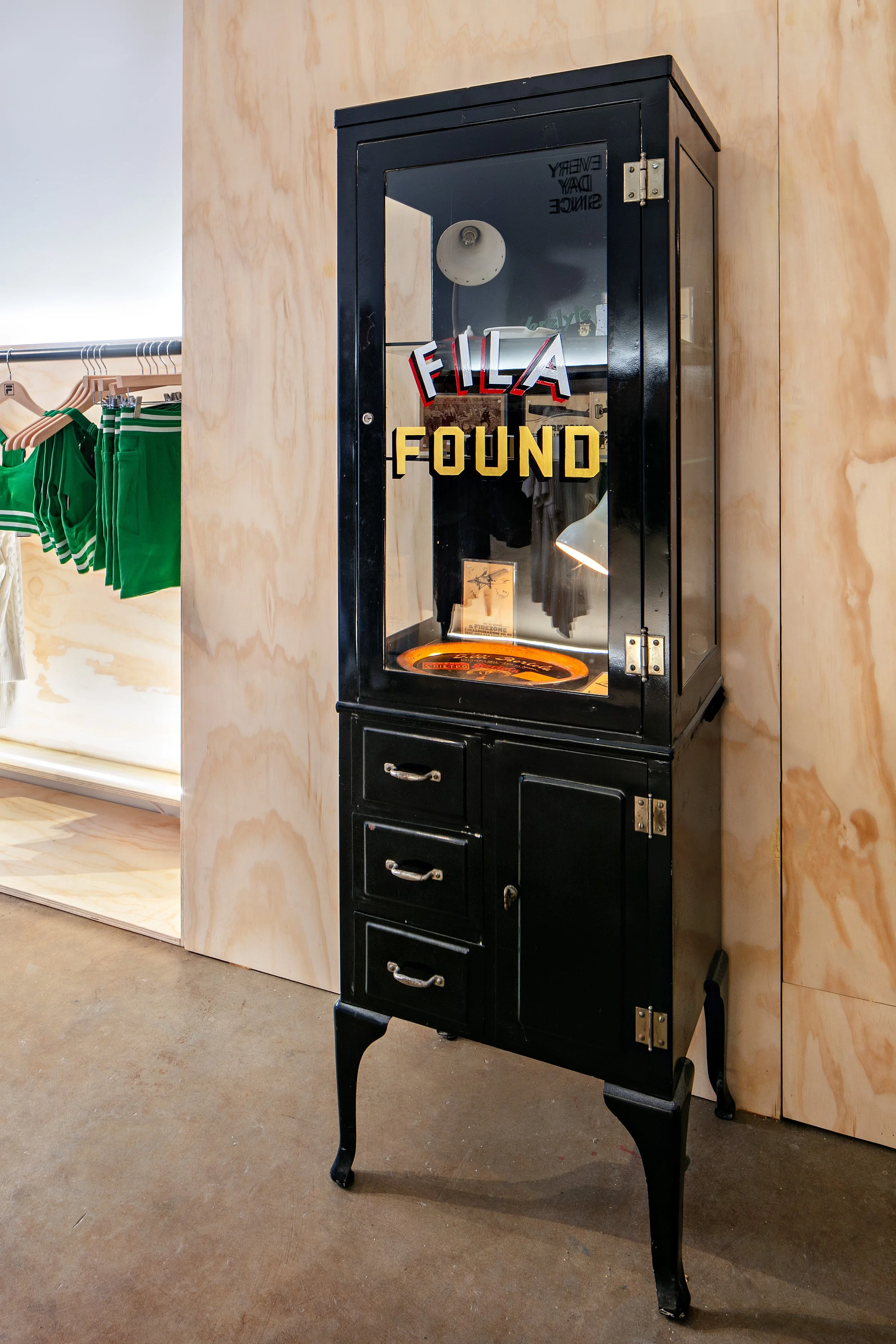 Vintage black display cabinet with "FILA FOUND" text, housing documents and a lamp, located in a store with wooden walls and clothing racks.