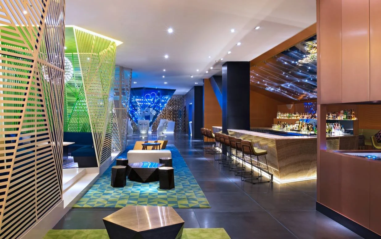 Modern hotel lounge featuring a stylish bar area with high stools, geometric patterned carpets, artistic lighting, and contemporary furniture.