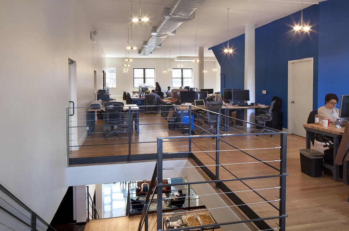Modern open-plan office with desks and computers, a blue accent wall, ceiling lights, and a staircase leading to a lower level.