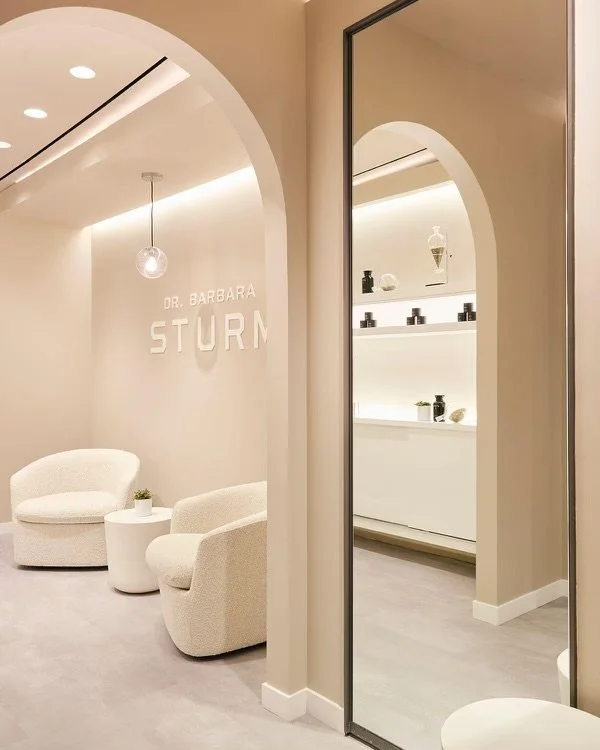 Minimalist waiting area with beige tones, featuring two cozy chairs, a small table, and a mirror. Wall displays 'Dr. Barbara Sturm' with shelves for products in the background.