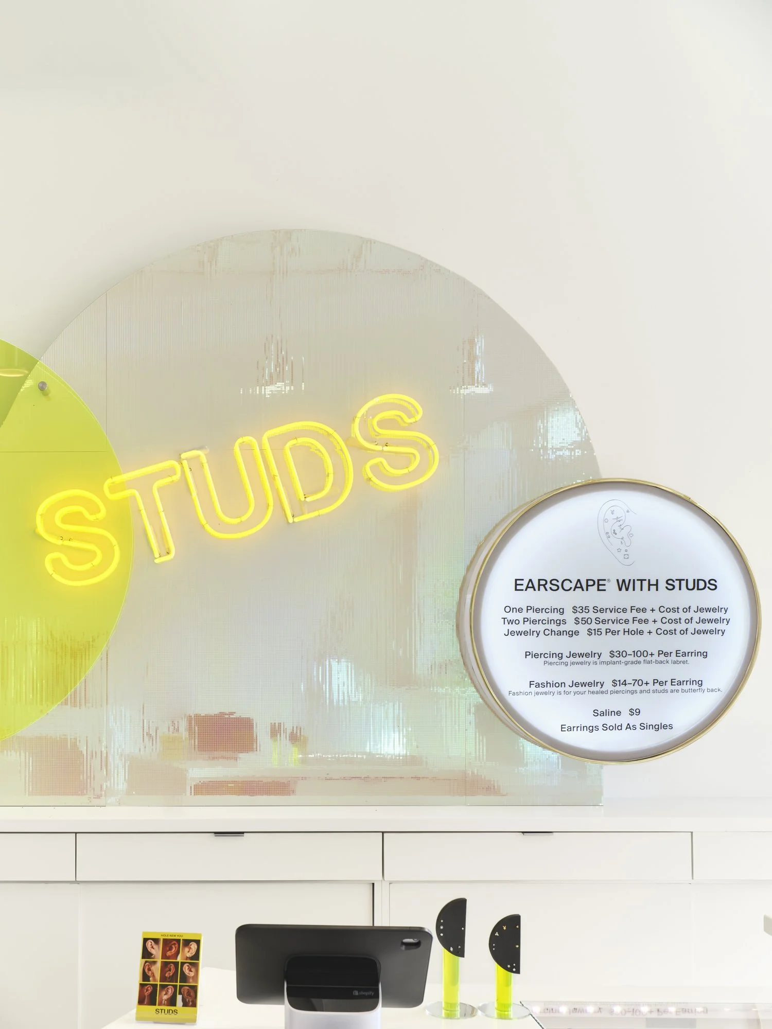 Store interior with neon 'STUDS' sign and pricing list for ear piercings and earrings, including service fees and jewelry costs, displayed on wall. Checkout area with payment terminal and display items.