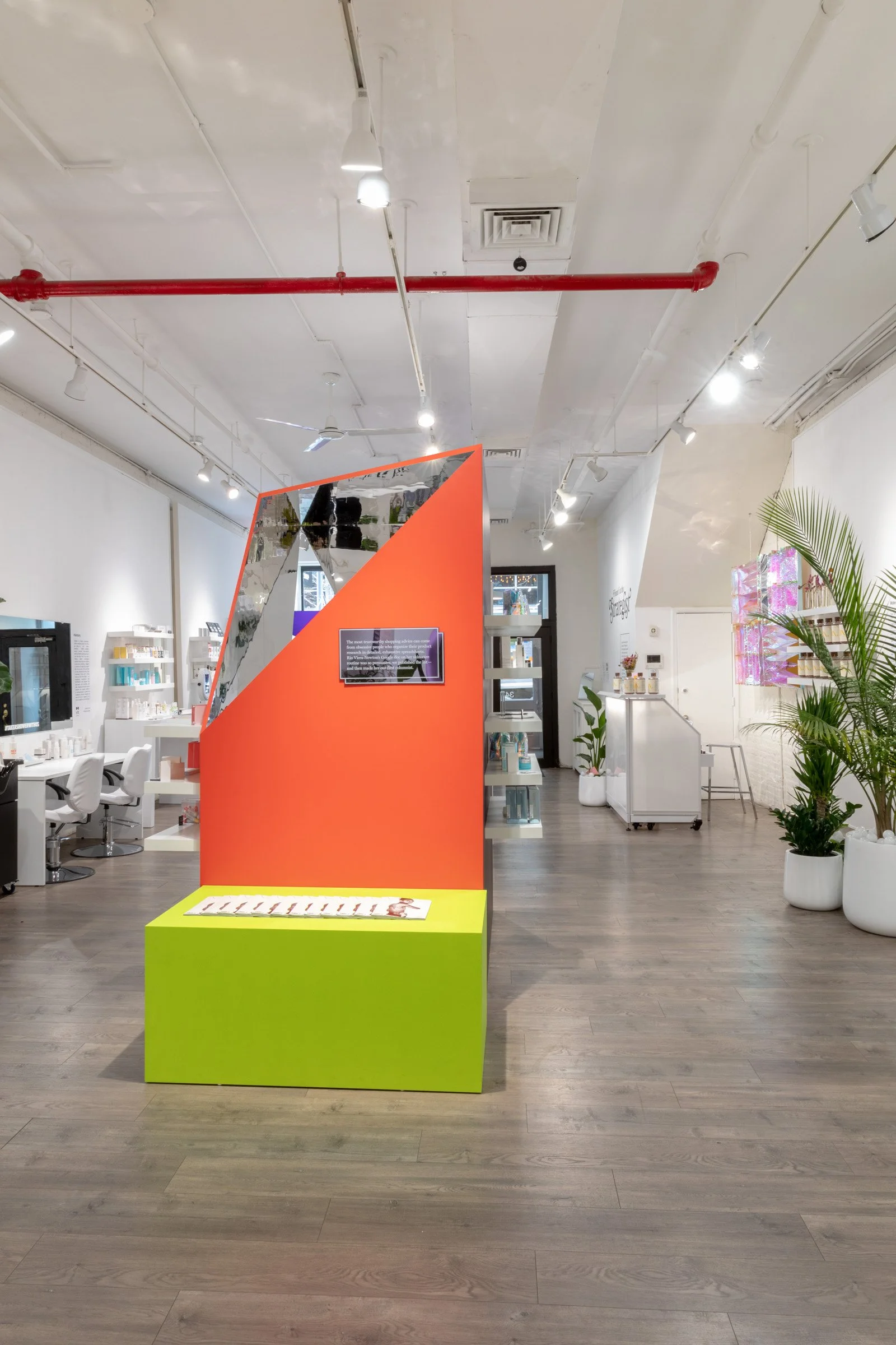 Interior of a modern salon with bright decor, featuring a large orange and green display structure in the center. The space has white walls, wooden floors, and decorative plants. Shelves with products are visible in the background along with salon chairs and lighting fixtures on the ceiling.