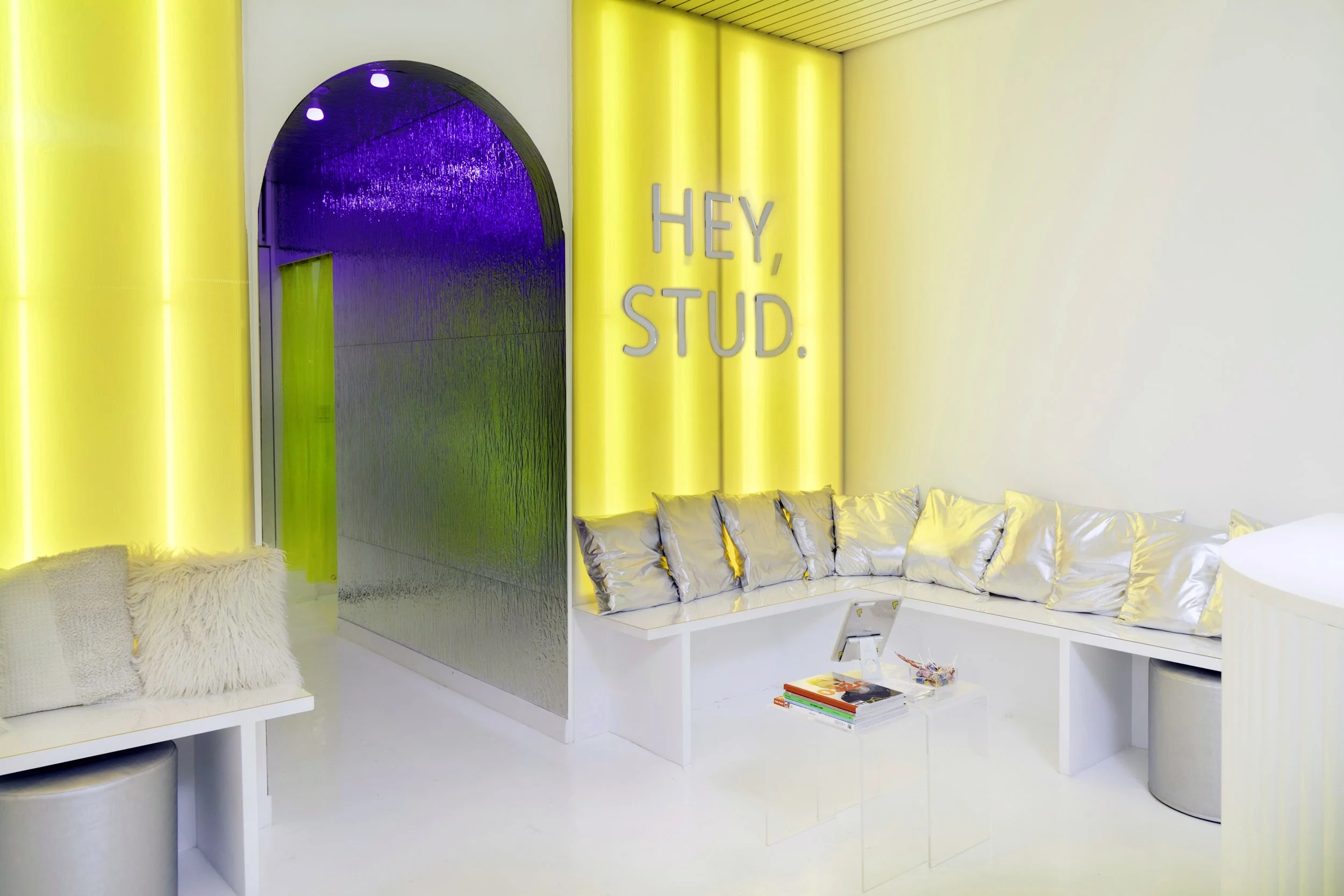 Modern lounge with yellow lighting, silver cushions, a clear table with books, and "Hey, Stud." on the wall.