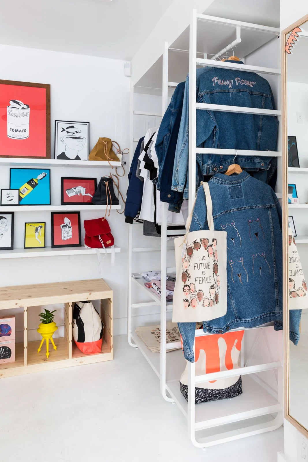 Boutique clothing store interior with denim jackets, framed art on walls, tote bags with feminist slogans, and small wood shelves holding bags and a plant.