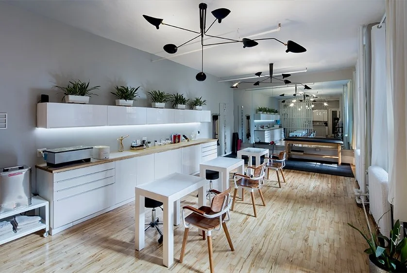 Modern nail salon interior with white furniture, wooden flooring, plants, large mirrors, and stylish black ceiling lights.