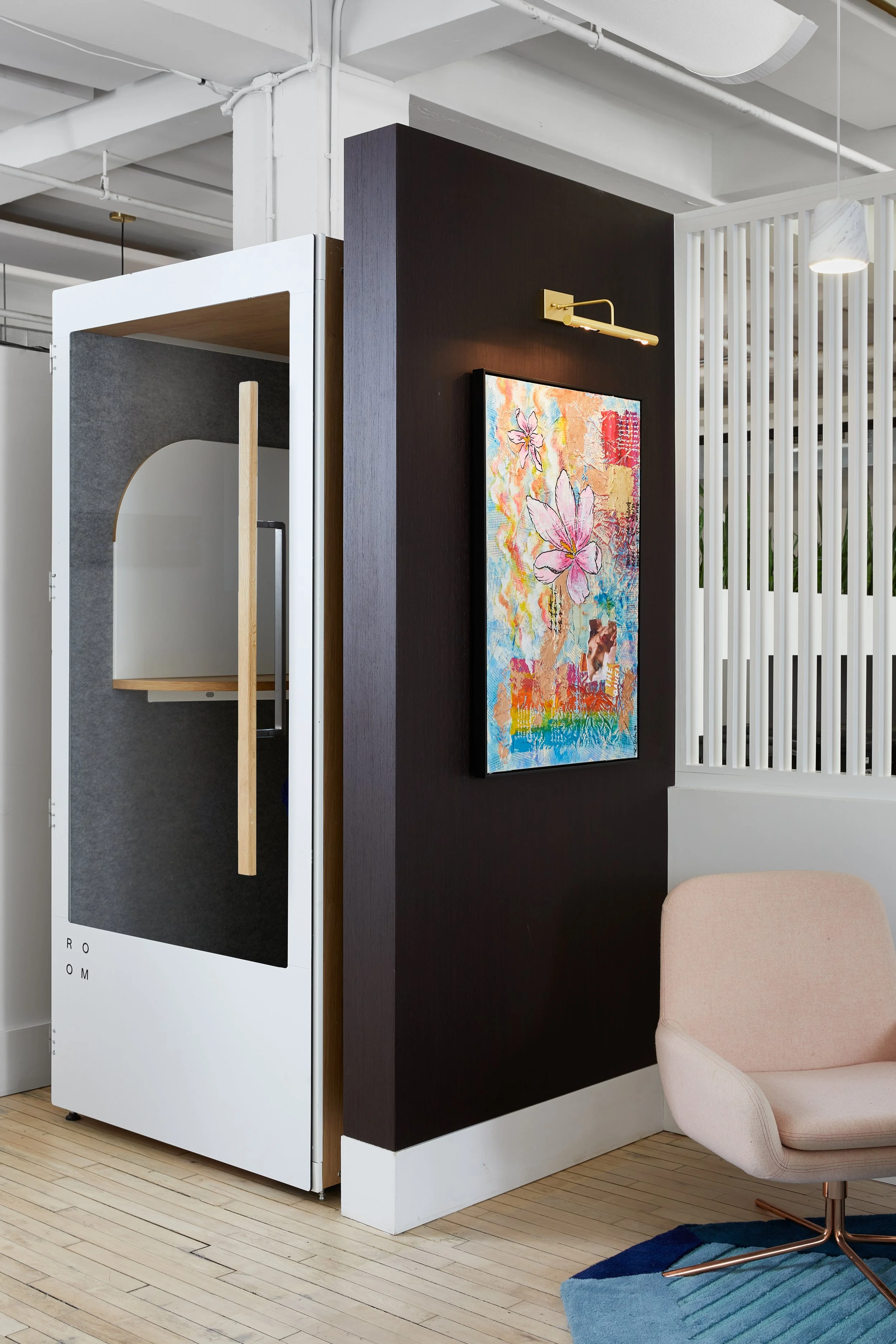 Modern office space featuring a small, enclosed phone booth with a wooden interior and glass door. A vibrant floral art piece is mounted on a dark wall, illuminated by a picture light. A pink swivel chair is positioned on a colorful geometric rug.