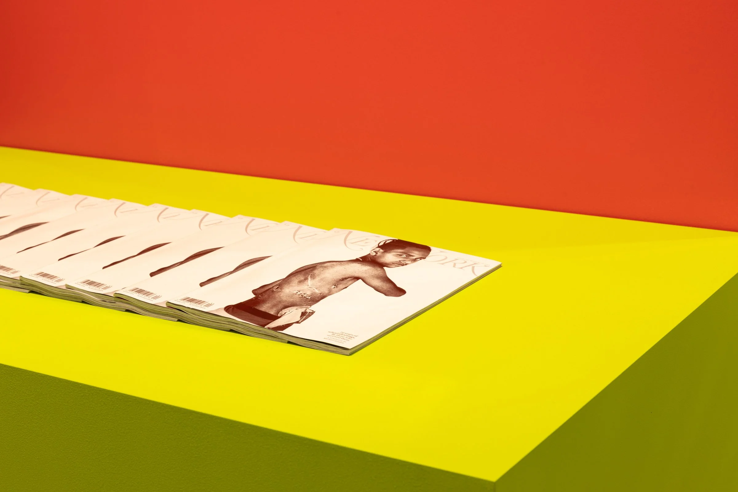 Stack of magazines with a cover featuring a shirtless man, on a bright yellow table against an orange background.