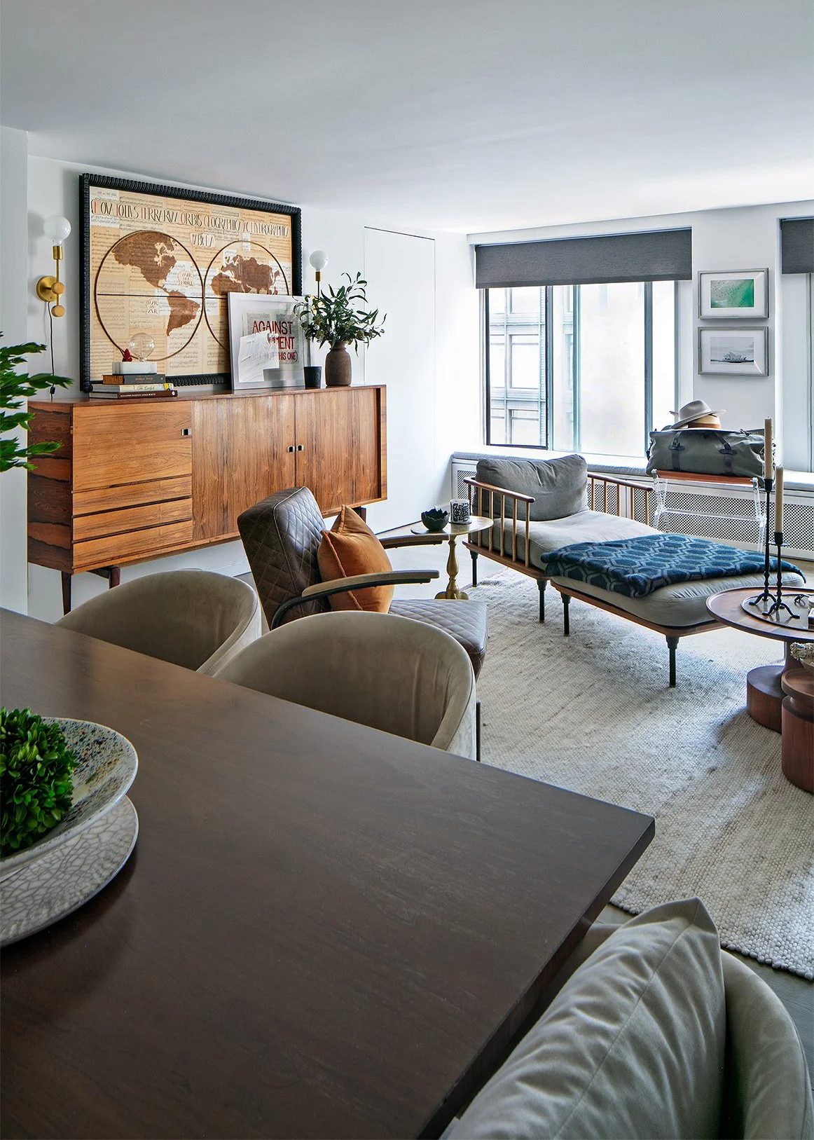 Modern living room with a wooden sideboard, world map art, plants, a lounge chair with a pillow, a daybed under the window, and framed pictures on the wall.