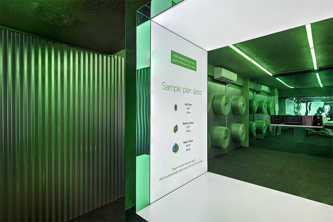 Interior of a green-lit storage facility with corrugated metal walls, shelving, and bubble wrap rolls. A sign displays "Sample plan sizes" with closet measurement options.