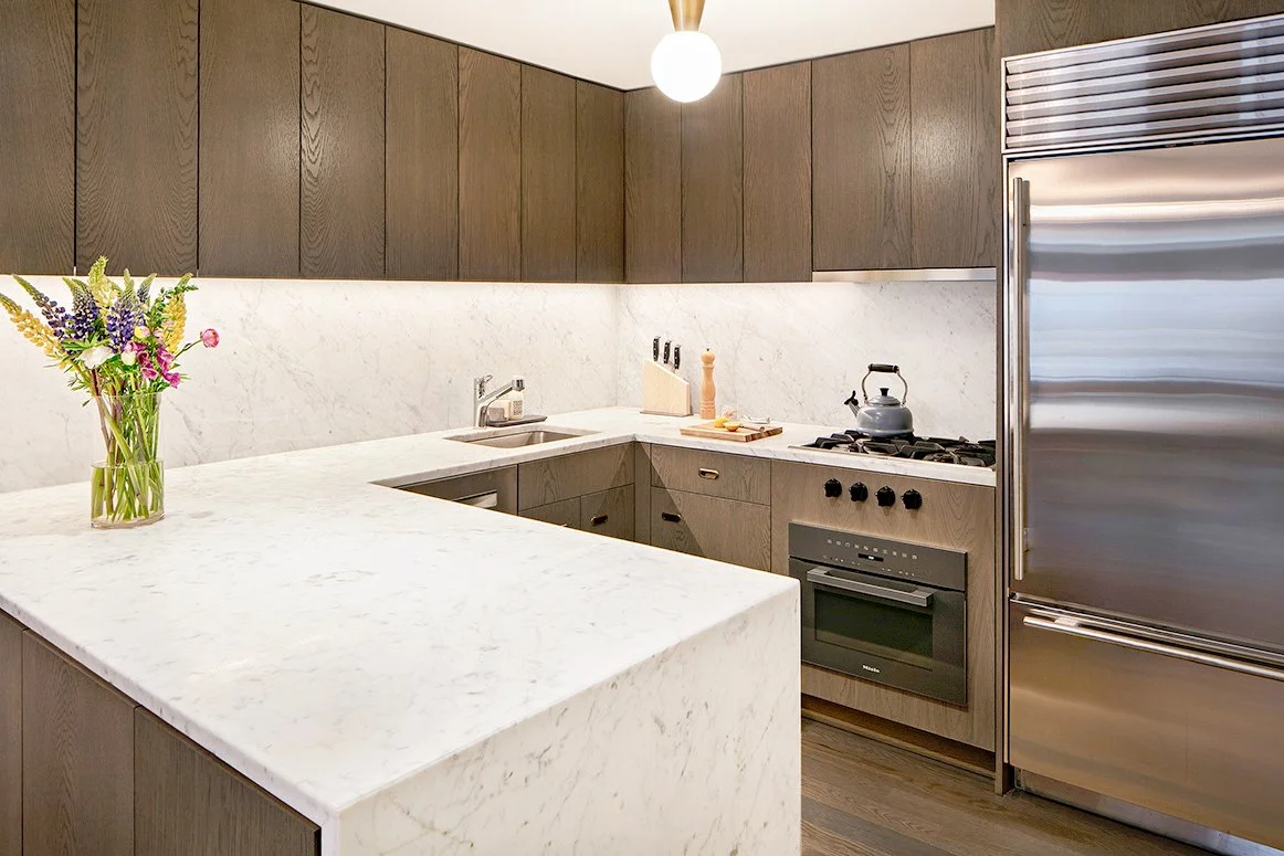 Modern kitchen with white marble countertops, dark wooden cabinets, a stainless steel refrigerator, a stovetop with a kettle, and a vase of colorful flowers.