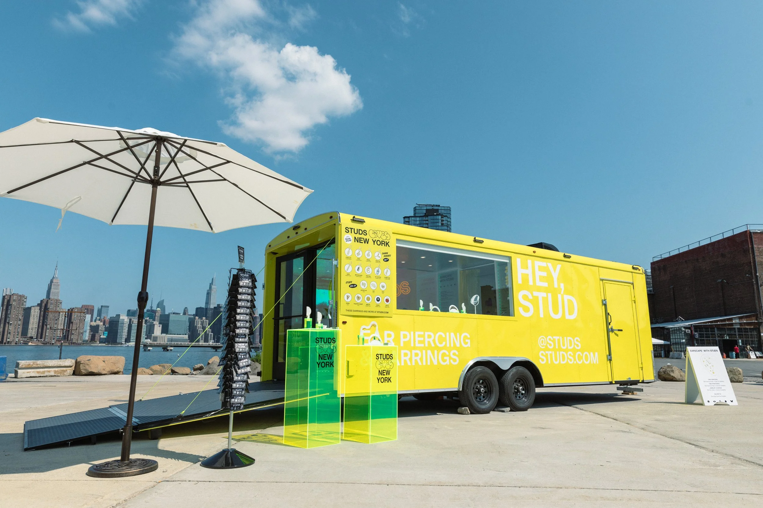 Yellow mobile truck with 'Hey, Stud' and 'Studs New York' written, offering ear piercing services, parked near a waterfront with a city skyline in the background, and an umbrella providing shade.
