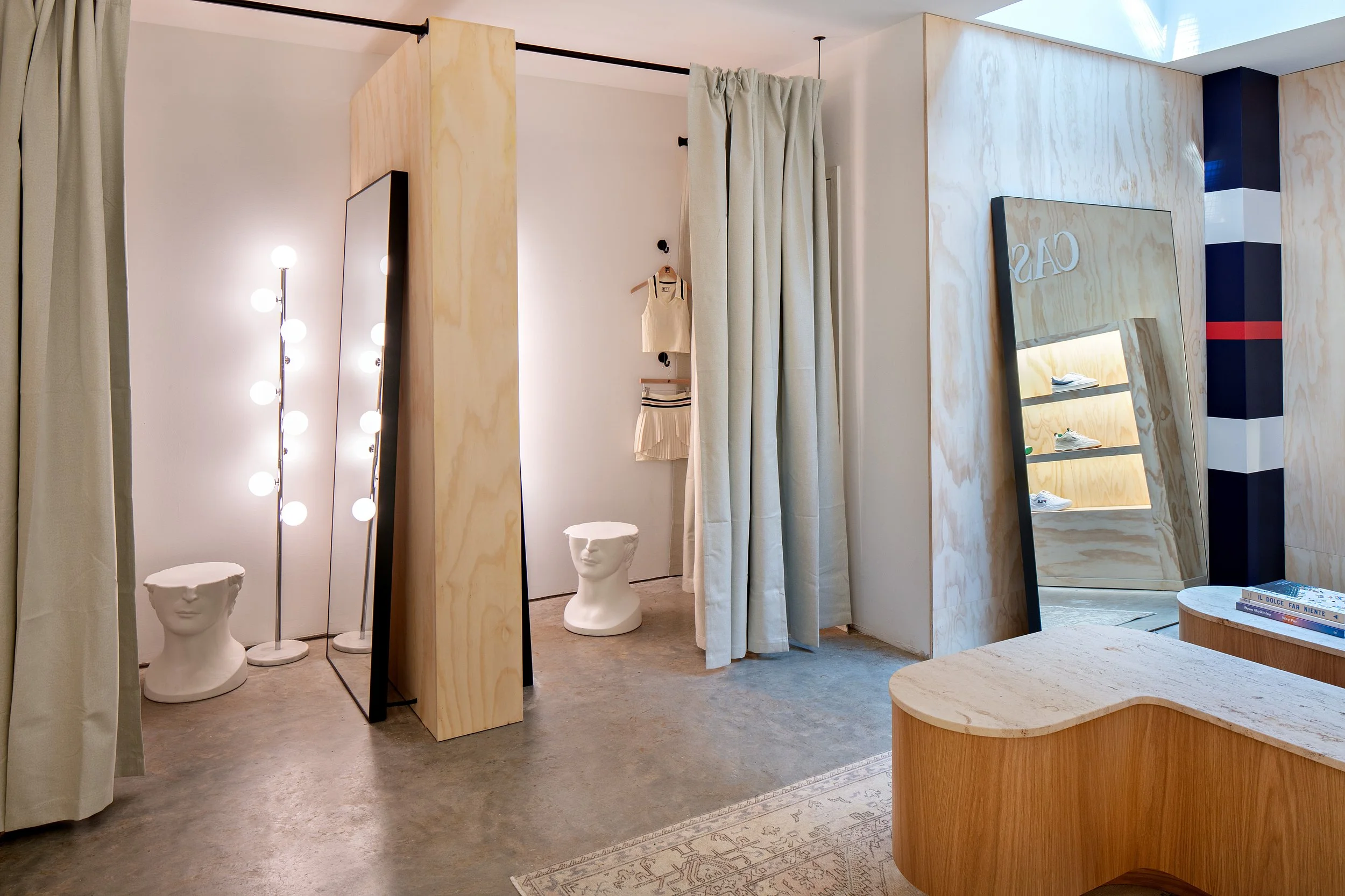 Modern fitting room with wooden accents, large mirrors, and soft lighting. A small display shelf with shoes is visible. Beige curtains and unique mannequin stools add elegance. Minimalist design with concrete flooring.