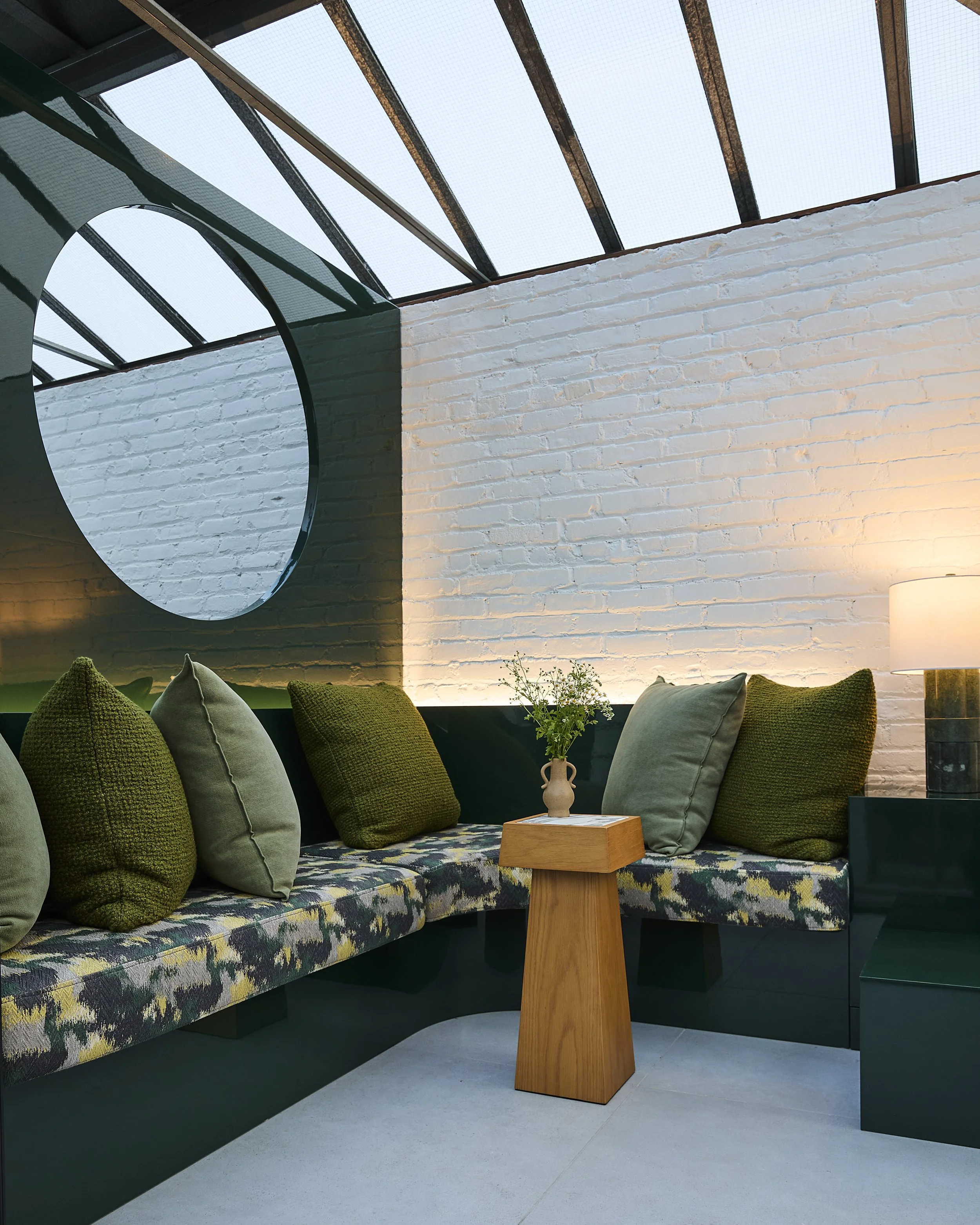 An interior sitting area with a built-in bench with multicolored upholstery and green and gray cushions, a wooden side table with a small vase of flowers, and a black side table with a lamp. The room has a white brick wall and a glass ceiling.