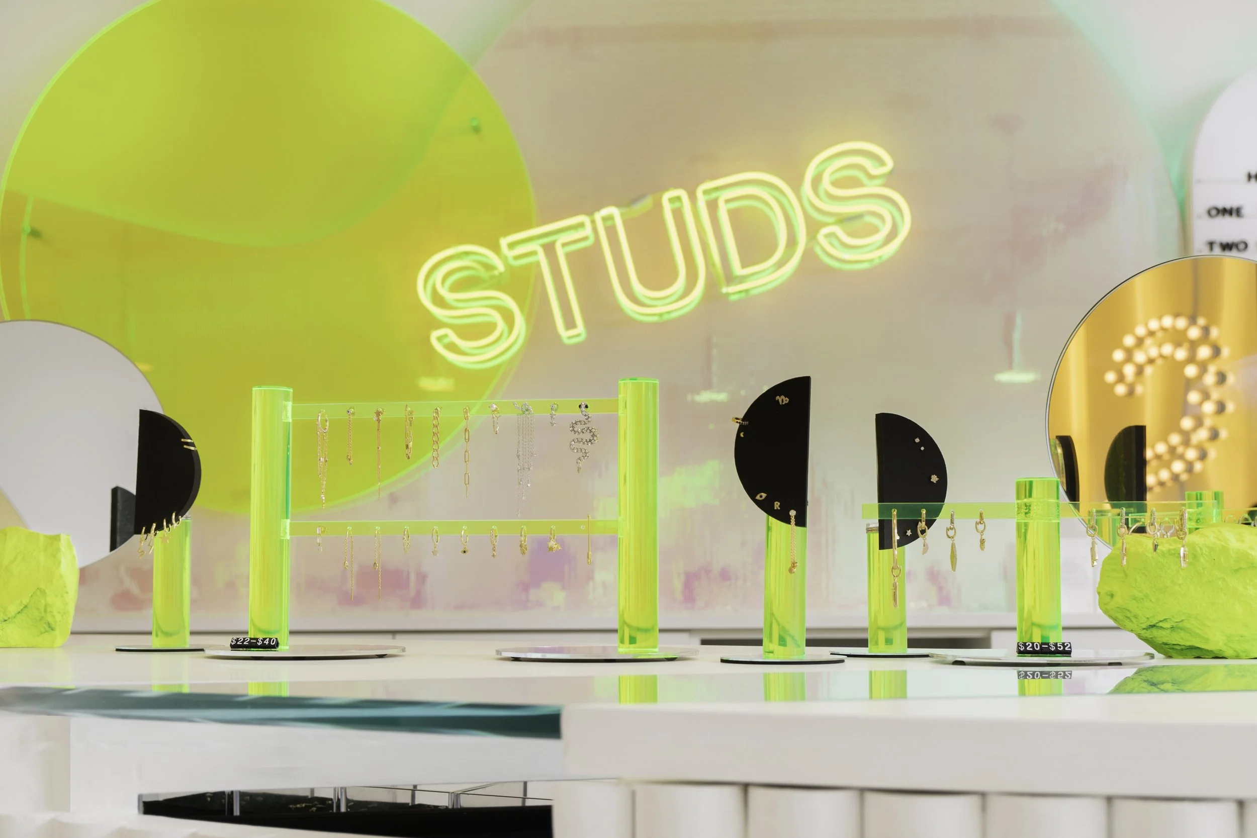 Jewelry store display with neon 'STUDS' sign, showcasing earrings on green acrylic stands with mirrors.