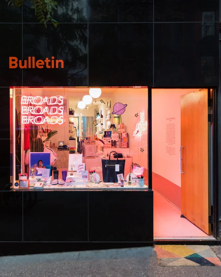Storefront with 'Bulletin' sign, showcasing various products and a neon 'BROADS' sign, with a view into a colorful interior.