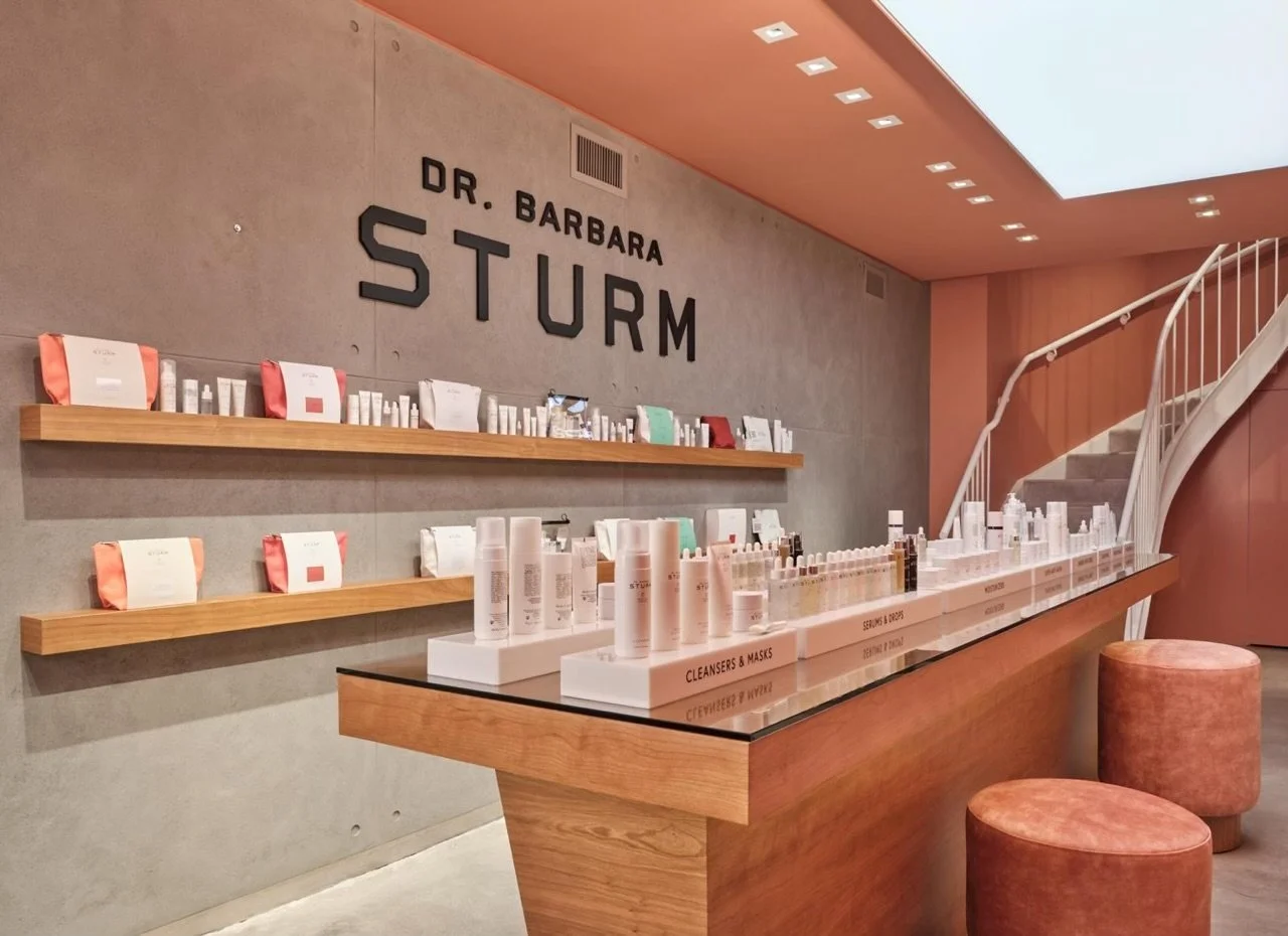 Cosmetics store interior with skincare products, shelves, and display racks, labeled Dr. Barbara Sturm. Modern design with wood accents, light fixtures, and spiral staircase.