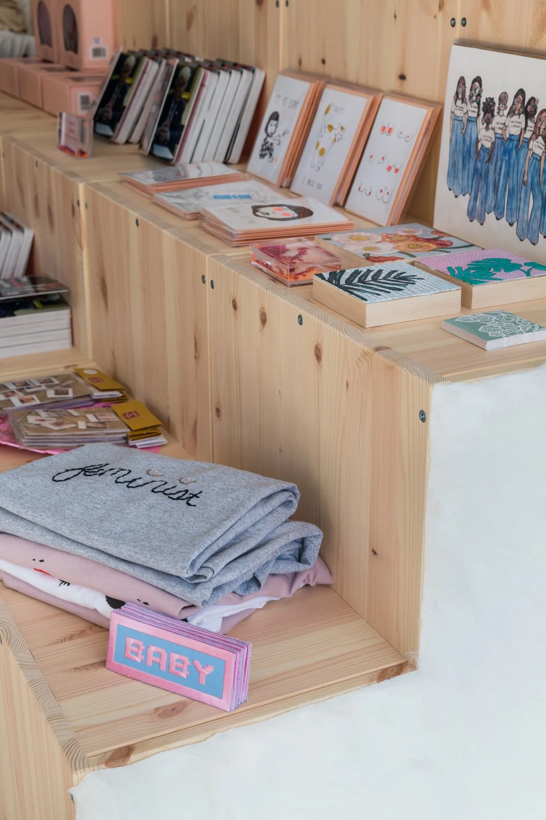 Wooden shelves displaying folded sweatshirts, art prints, and books, including a sweatshirt with 'feminist' text and a sign saying 'BABY.'