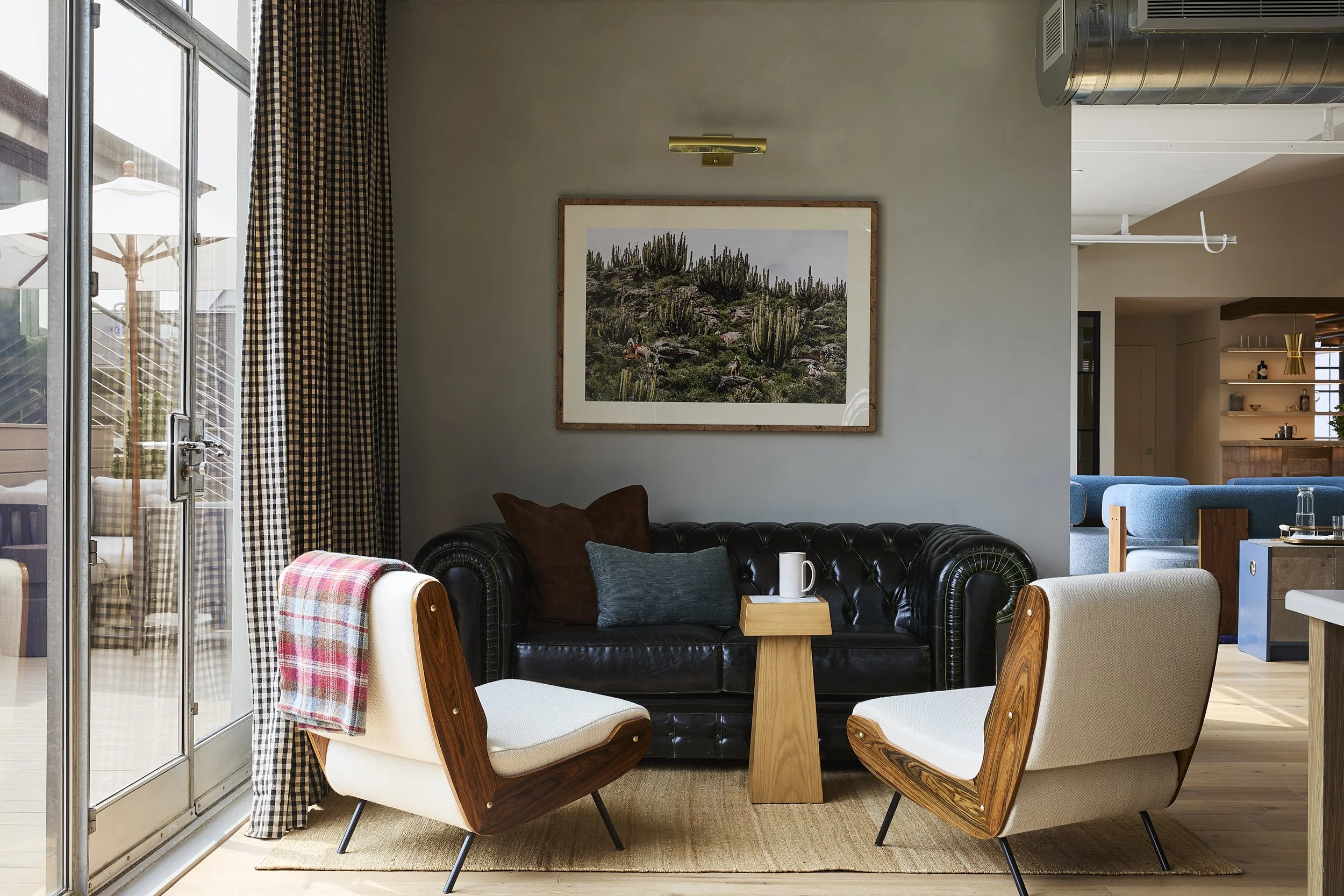 Living room with a black leather sofa, two mid-century modern armchairs, a small wooden side table with a mug and a book, a framed landscape photo of cacti on the wall, and sliding glass door with gingham curtains leading to an outdoor patio with patio furniture and umbrellas.