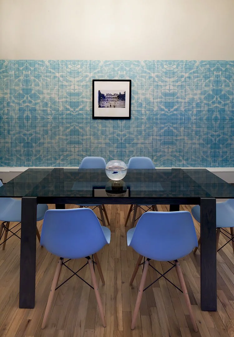 Modern dining room with black glass table, blue chairs, and blue patterned wall with a framed photo.