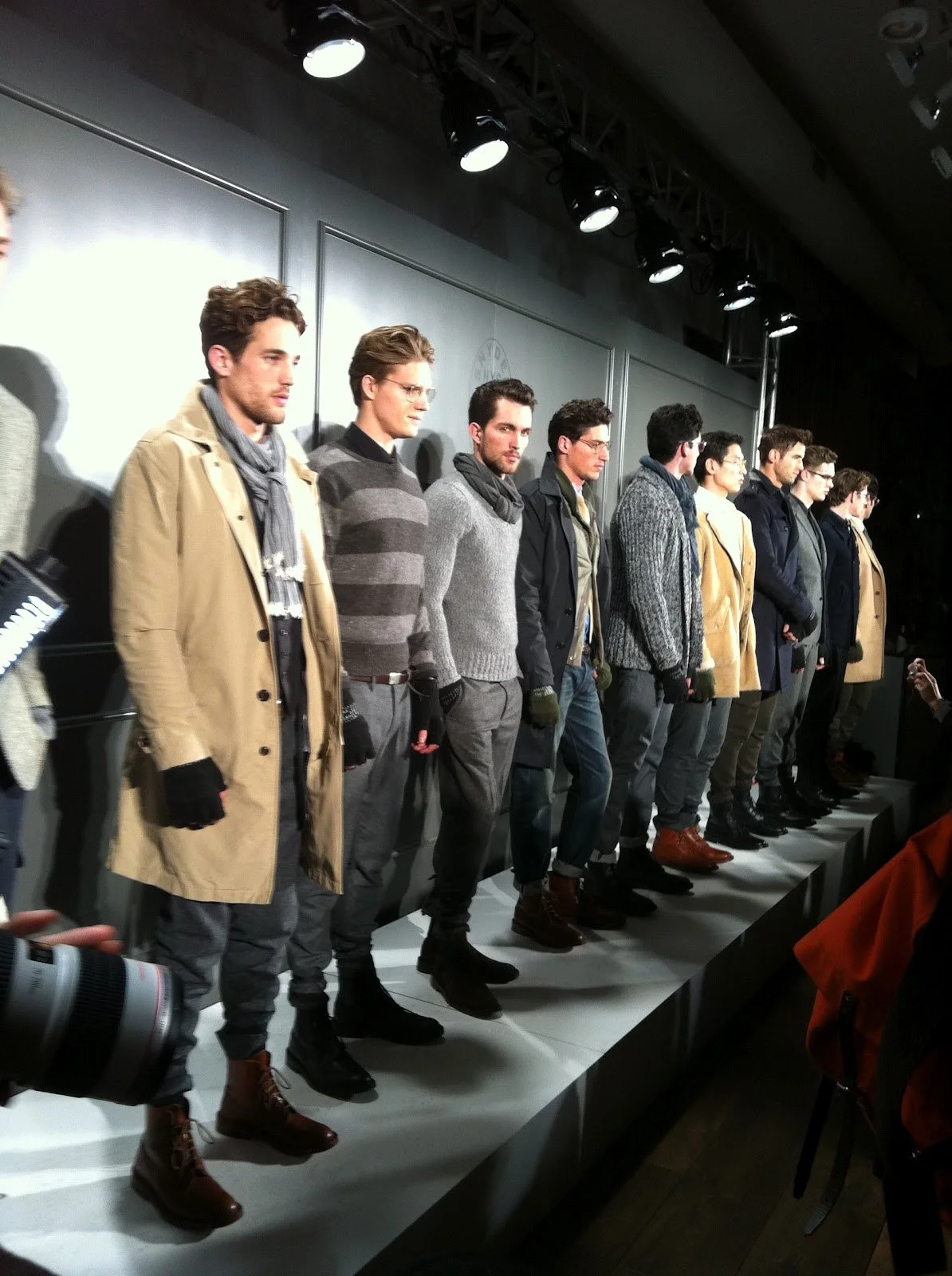 Group of male models in winter fashion, including coats and knitwear, standing on a runway under bright lighting at a fashion event.