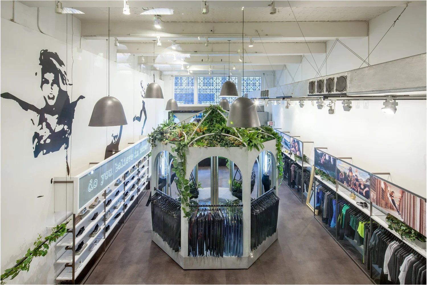 Interior of a modern clothing store with wall art, green plants, and displays of shoes and clothes. Large pendant lights hang from the ceiling, and the store features a central display rack with hanging garments.