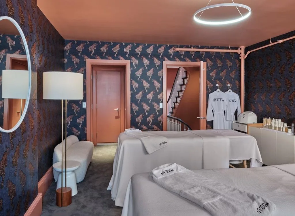 Interior of a spa treatment room with two massage tables covered in white linens, decorated with a leopard print wallpaper. A circular mirror and floor lamp are beside a small upholstered bench. Two robes hang on a wall, and skincare products are displayed on a cabinet.