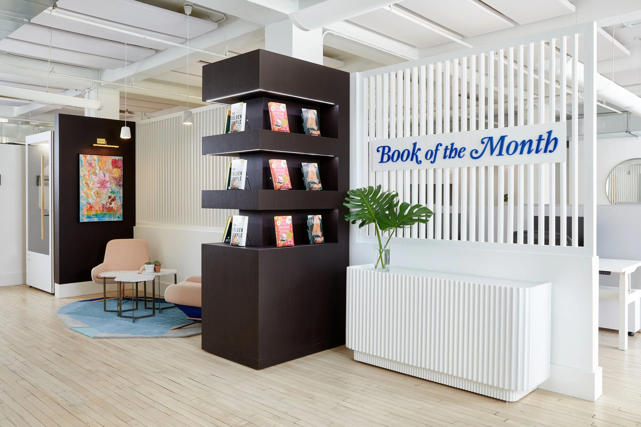Modern office interior with a book display labeled 'Book of the Month', featuring books on shelves, pink chairs, a small round table, palm leaves in a vase, and abstract wall art.