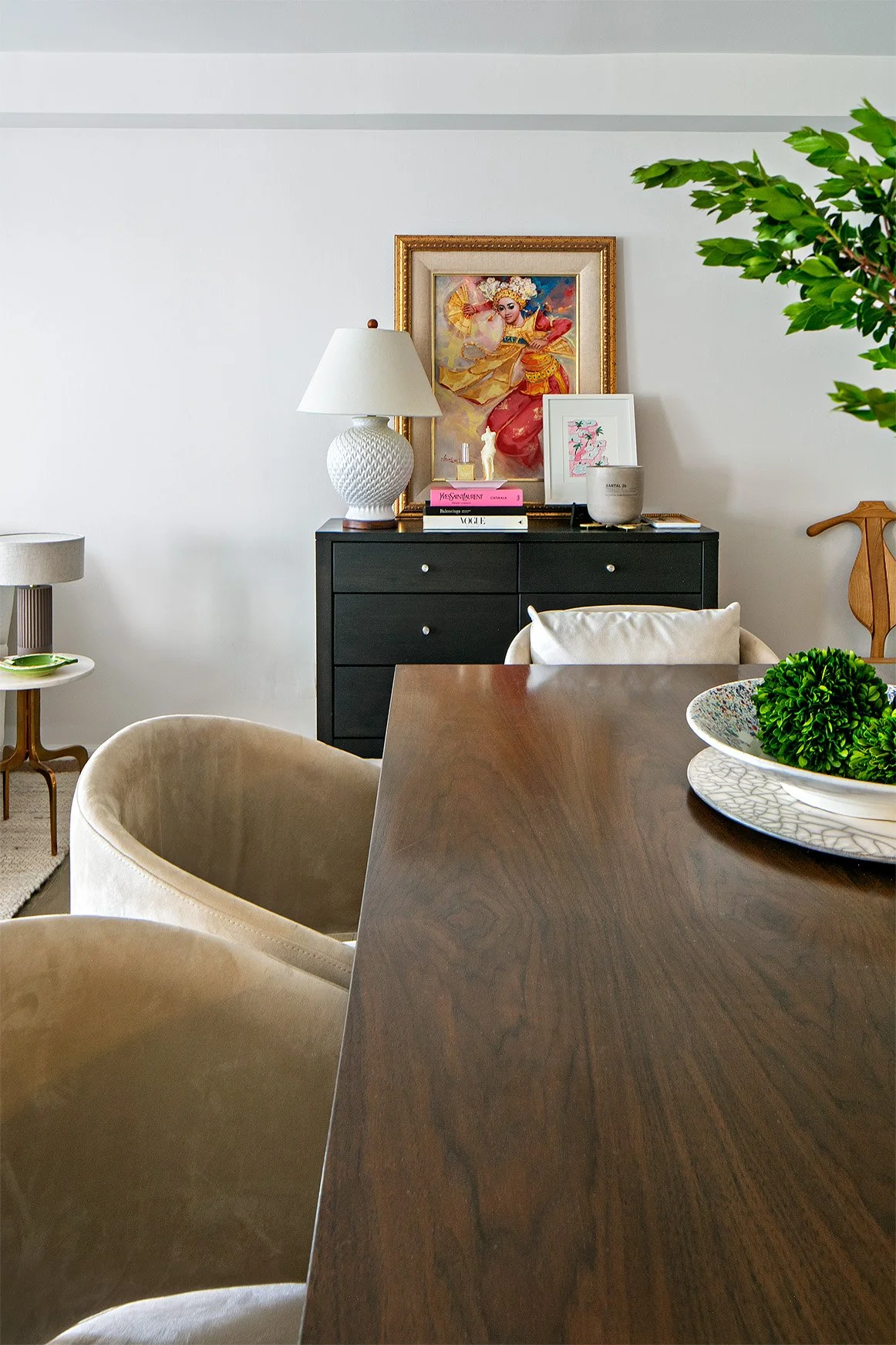 Interior living room with a wooden dining table, beige upholstered chairs, a black sideboard with a white lamp, framed artwork, books, and decorative items, and a large green potted plant.