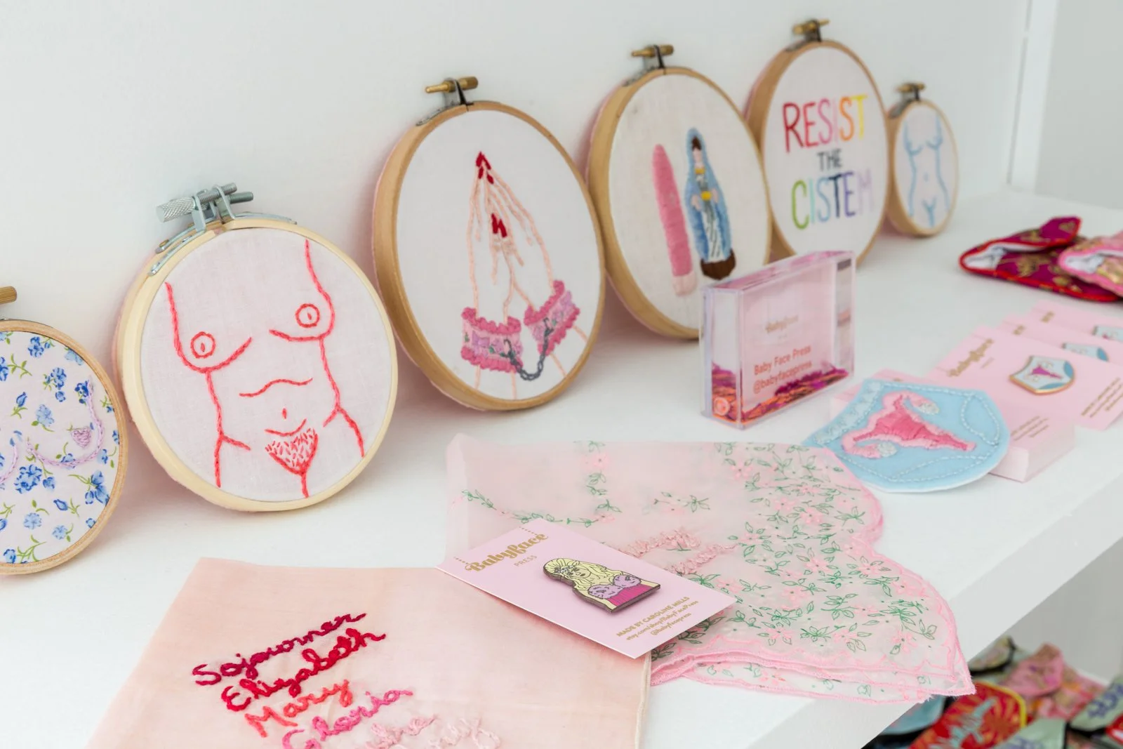 Display of various embroidered art pieces, including feminist themes, displayed with textiles and pink promotional material on a white surface.