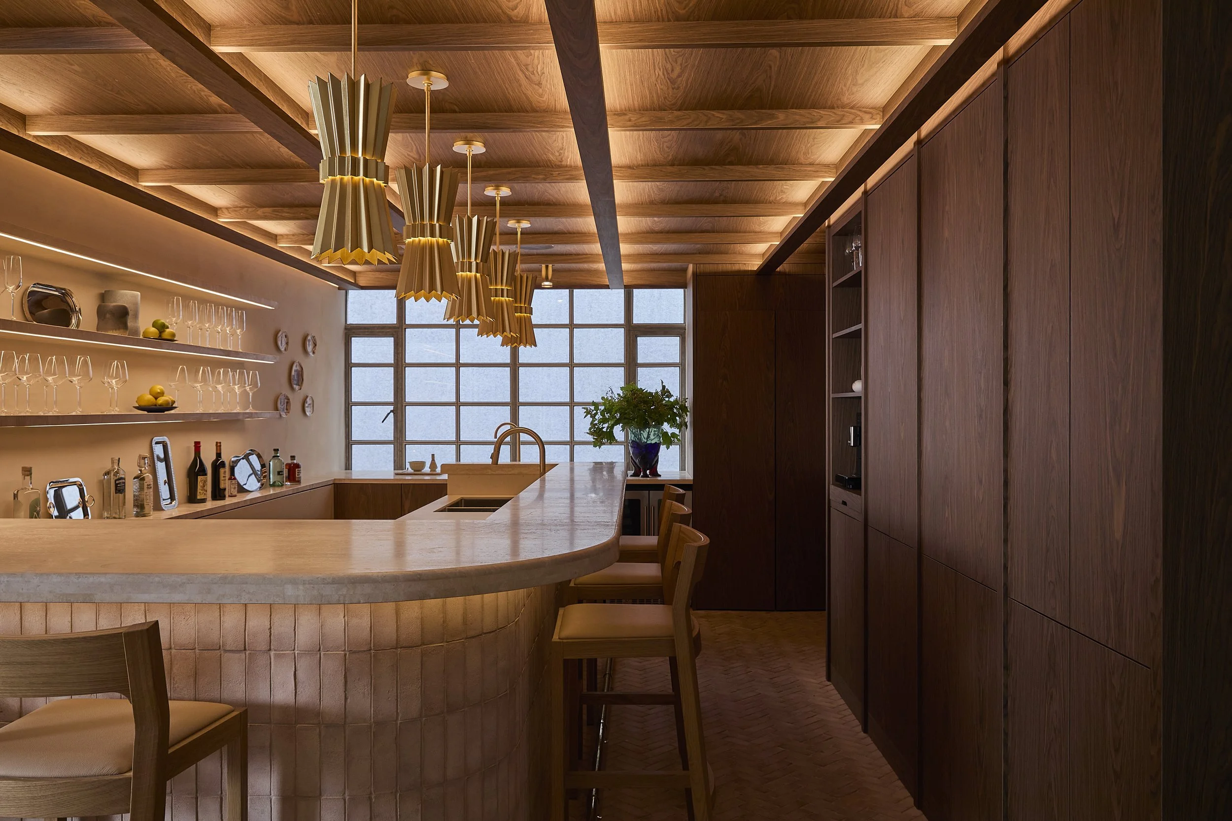 Interior view of a modern kitchen with wooden cabinetry, a curved island counter, pendant lights with paper shades, shelves with glassware and bottles, and a window at the back with a potted plant.