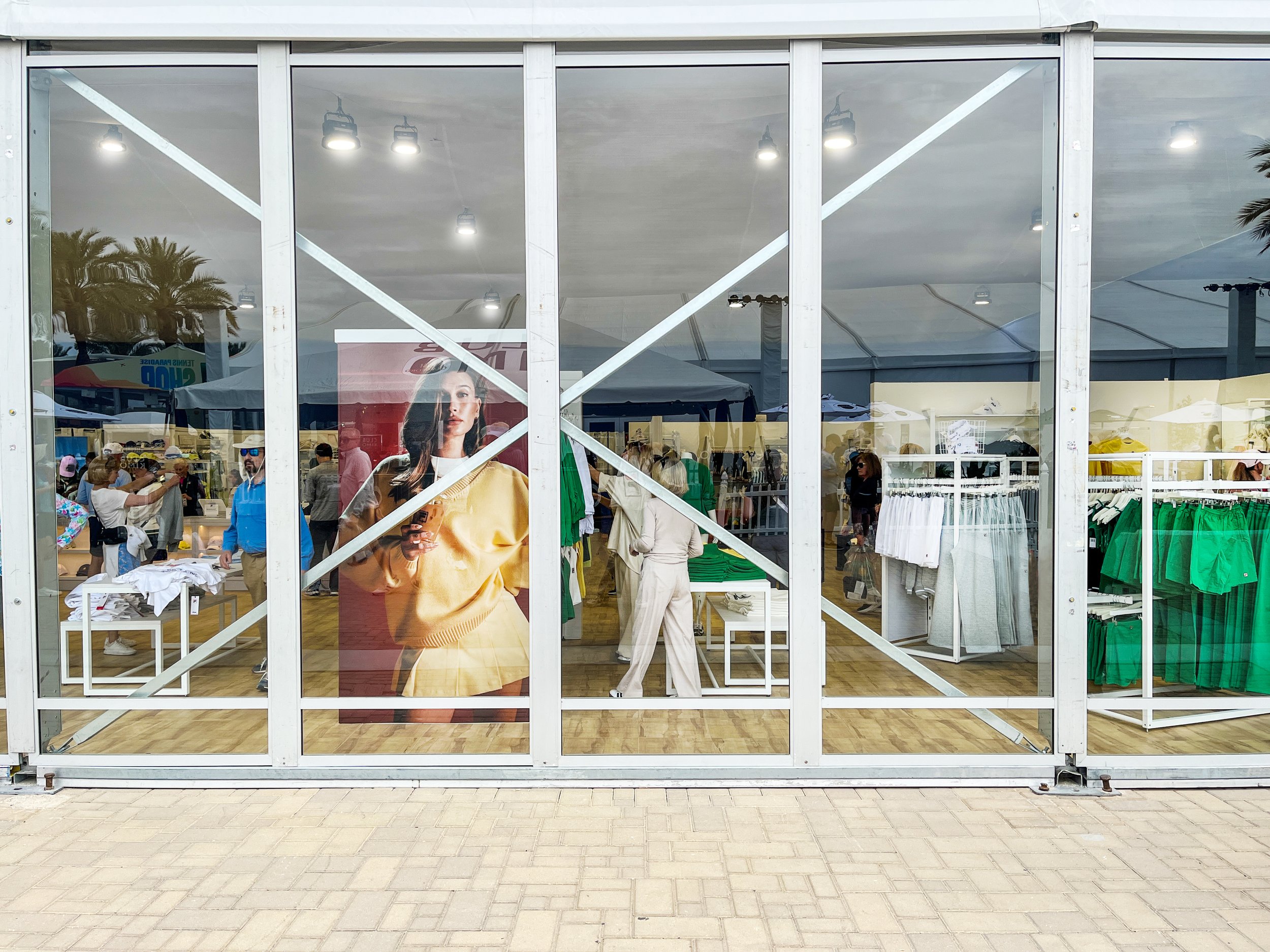 Clothing store with glass facade, displaying colorful garments, including green skirts and white shirts. Large promotional poster features a woman in a yellow outfit. Indoor lighting and shoppers are visible inside.