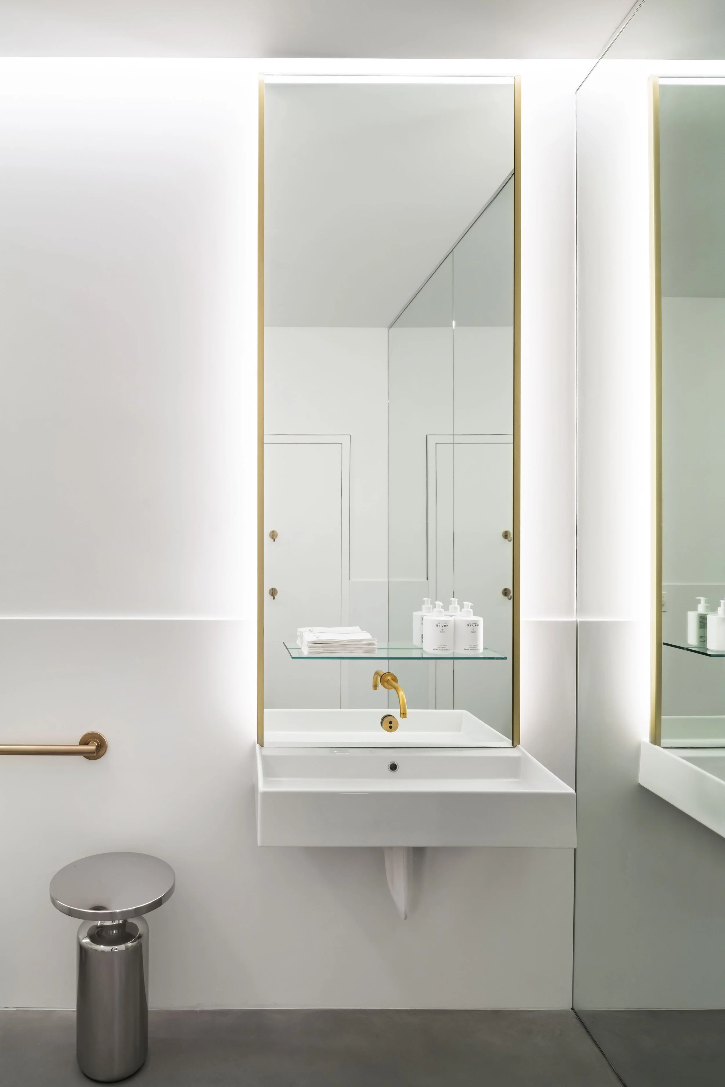 Modern bathroom with a white sink, gold faucet, wall mirror, and metallic stool. A glass shelf holds white bottles and folded towels. The room features minimalist design with clean lines and neutral colors.