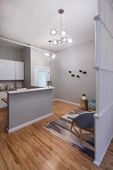 Modern minimalist living room and kitchen with light wood flooring, a small gray rug, a gray and white color scheme, and contemporary light fixtures. It includes a small seating area with a chair and a side table, and a partially visible kitchen with cabinets and a countertop.