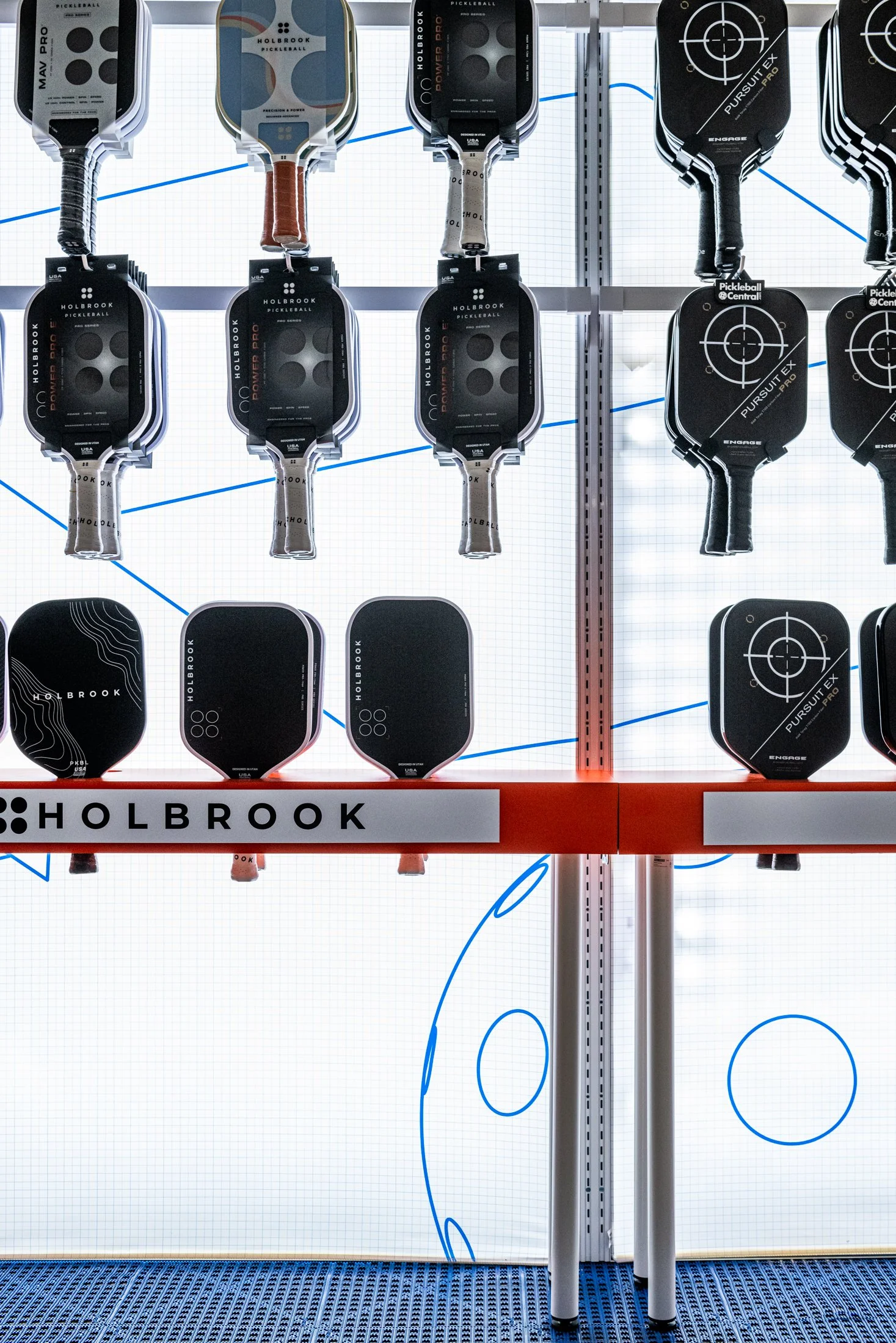 Display stand with various pickleball paddles, featuring Holbrook and Pursuit EX brands, in a store or showroom.