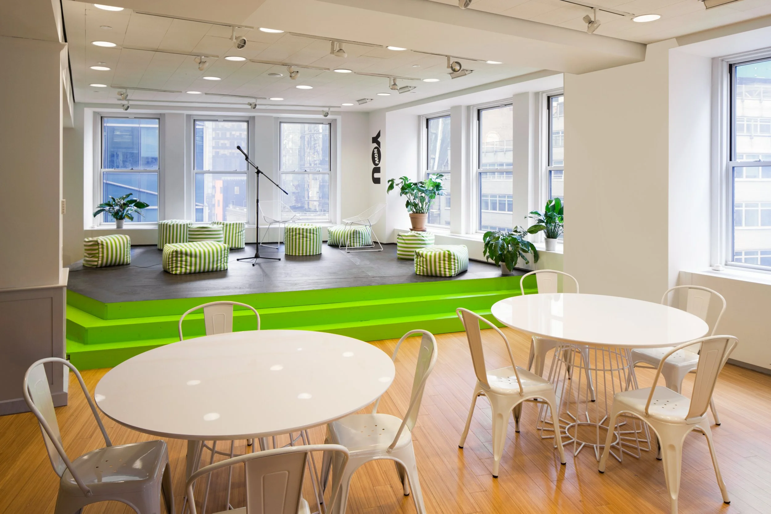 Modern office space with stage and seating area, featuring green striped pouffes, a microphone on a stand, and white chairs around round tables. Large windows and potted plants embellish the room.