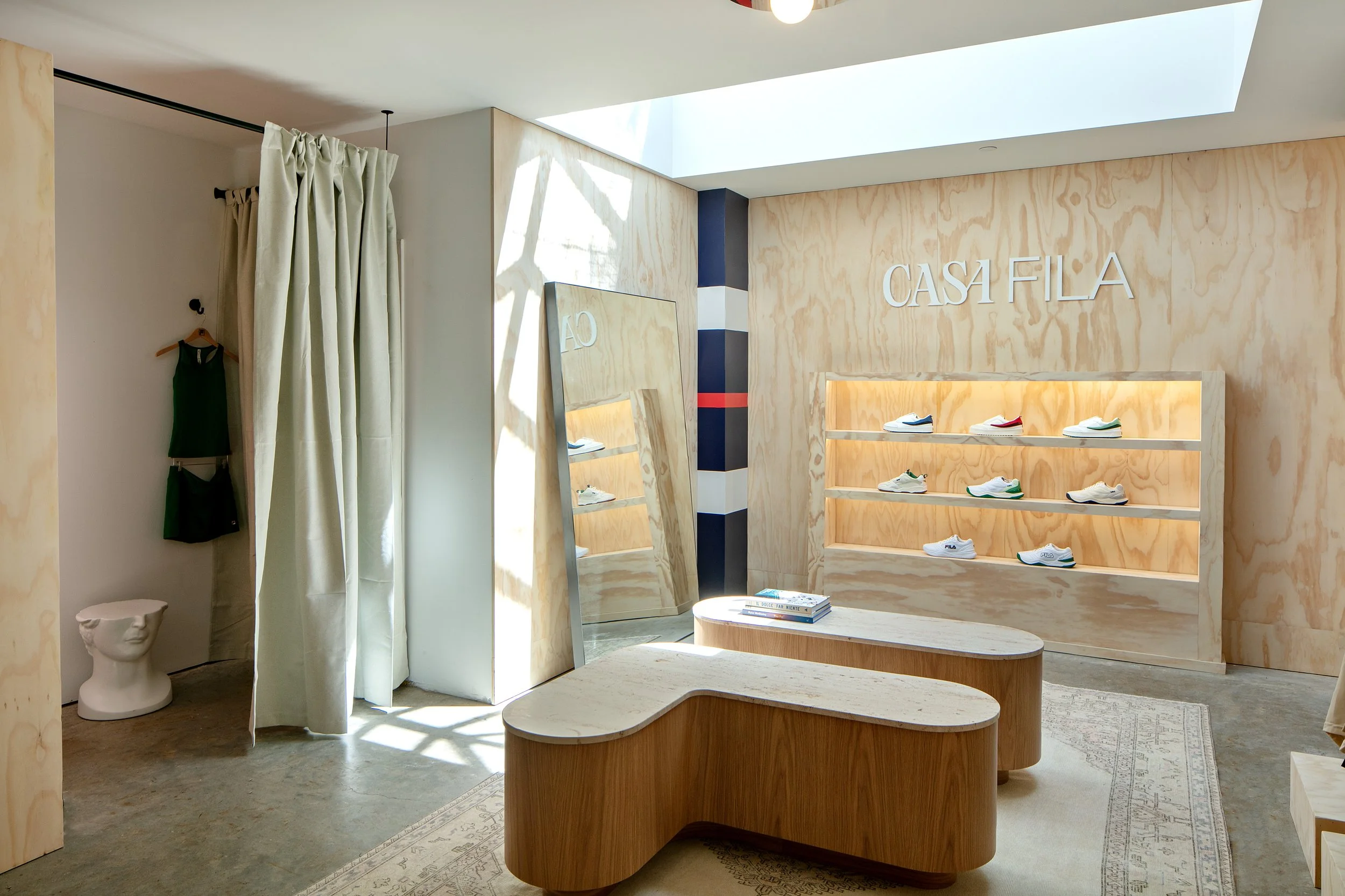Interior of a modern shoe store with wooden shelves displaying sneakers and a large mirror reflecting the decor. There's a changing area with a curtain on the left and a unique sculpture on the floor next to a hanging green garment. "Casa Fila" is written on the wall above the shoe display.