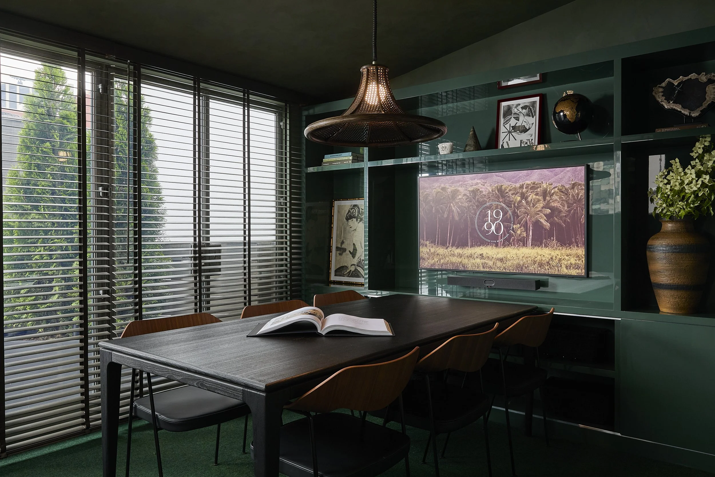 Modern dining room with a dark wooden table, six chairs, an open book, large window with horizontal blinds, green carpet, wall-mounted flat-screen TV displaying a landscape, decorative black globe, framed artwork, potted plant, and a hanging light fixture.